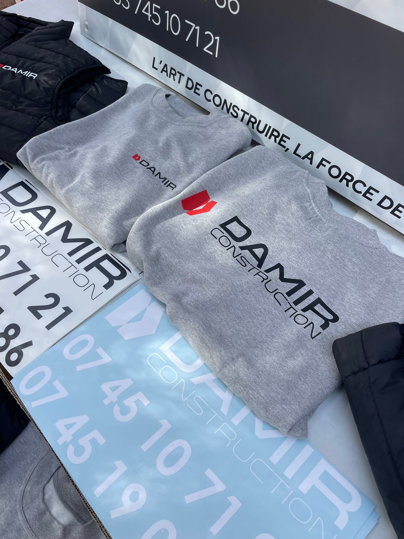 Vêtements, une veste noire, un pull gris et un sweat gris avec le logo DAMIR, exposés sur une table. Des affiches et des panneaux avec des numéros et des textes en français sont également visibles.
