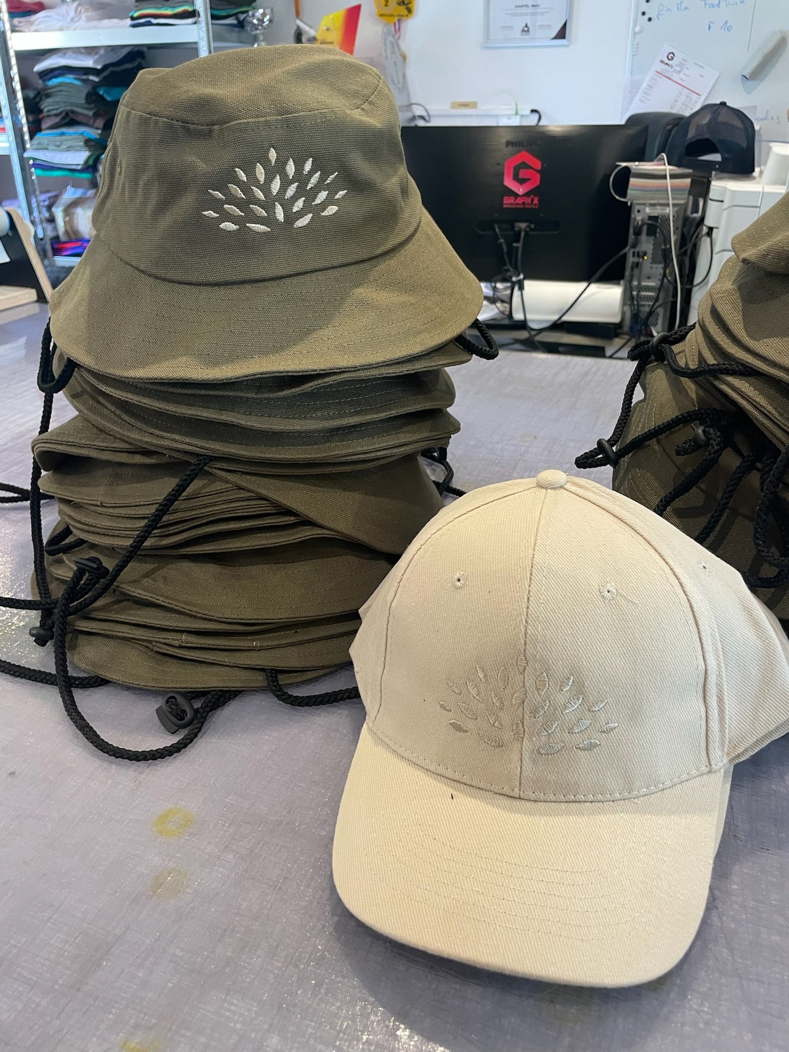 Une pile de casquettes de couleur beige avec un trou en forme de motif de feuilles, placée sur une surface de travail. En arrière-plan, un espace de bureau avec un ordinateur, des étagères et des papiers.