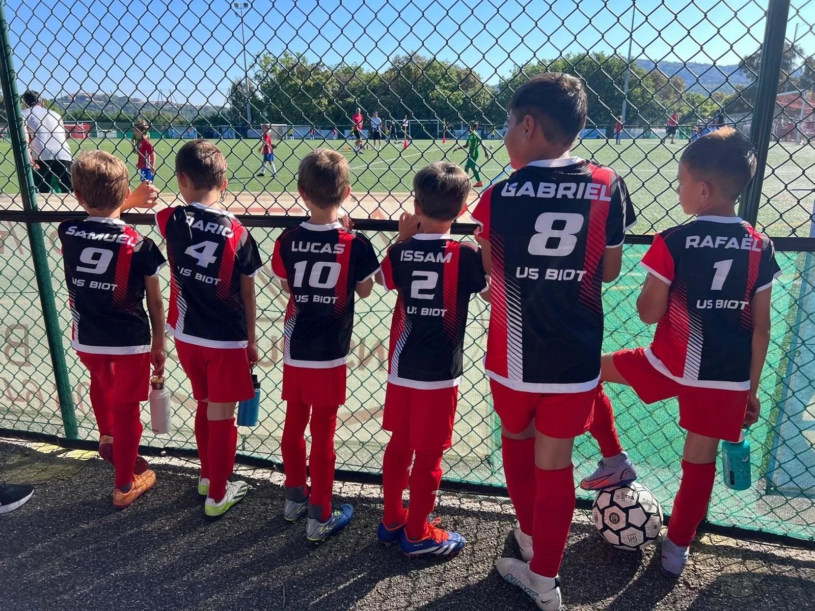 Jeunes joueurs de football uniformes noirs et rouges regardant un match depuis la clôture.