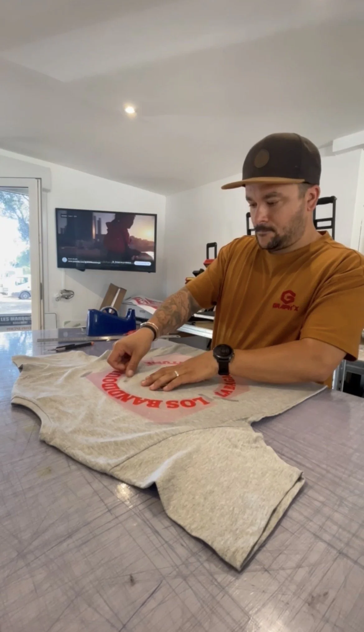 Un homme pose une affiche sur une table en travaillant avec ses mains, portant un t-shirt orange, une montre et un chapeau gris avec une visière beige.