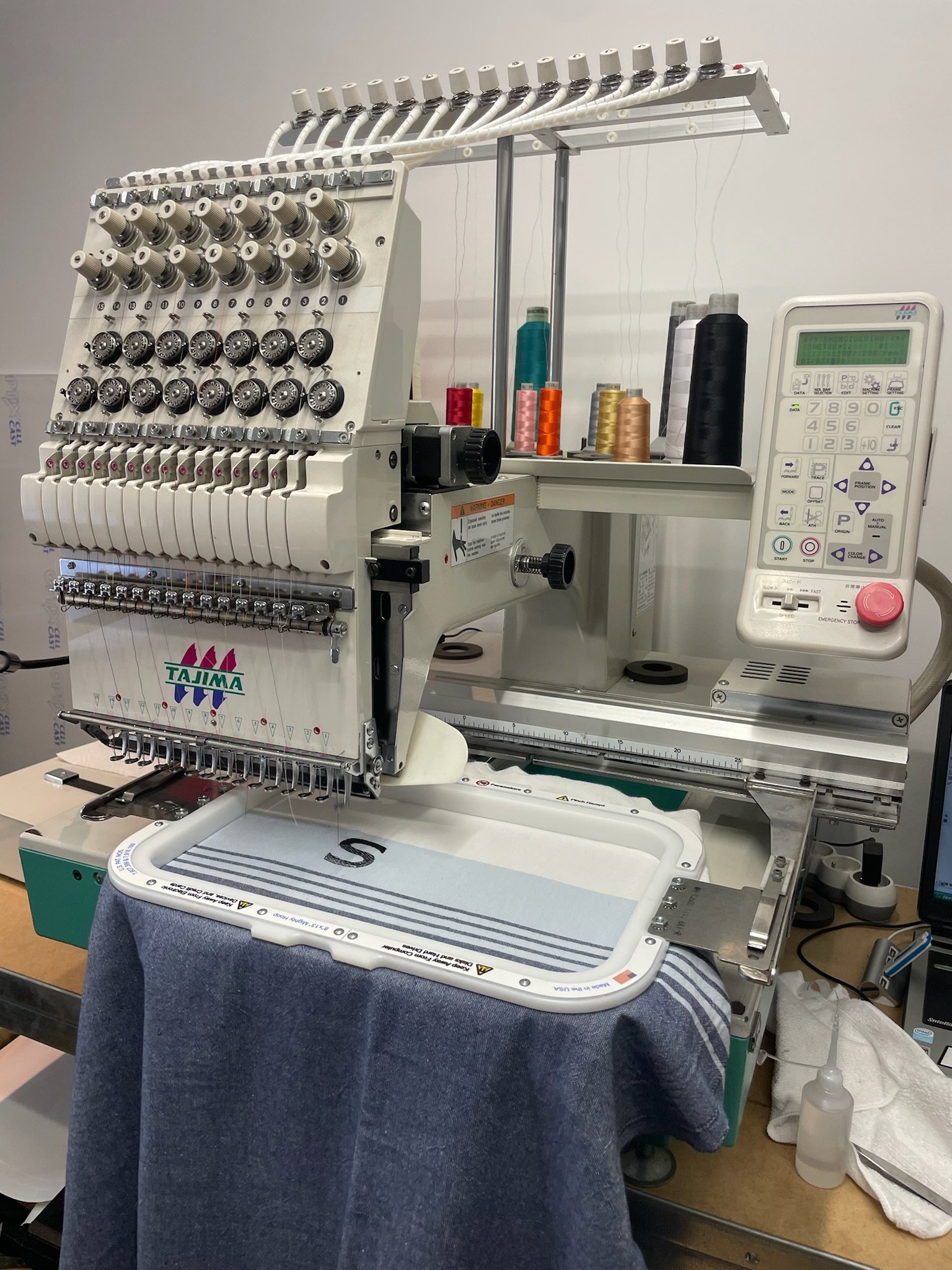 Une machine à broder en atelier, avec un cadre de broderie blanc, une personne en jeans en train de travailler dessus, entourée d'outils et de fournitures de couture.