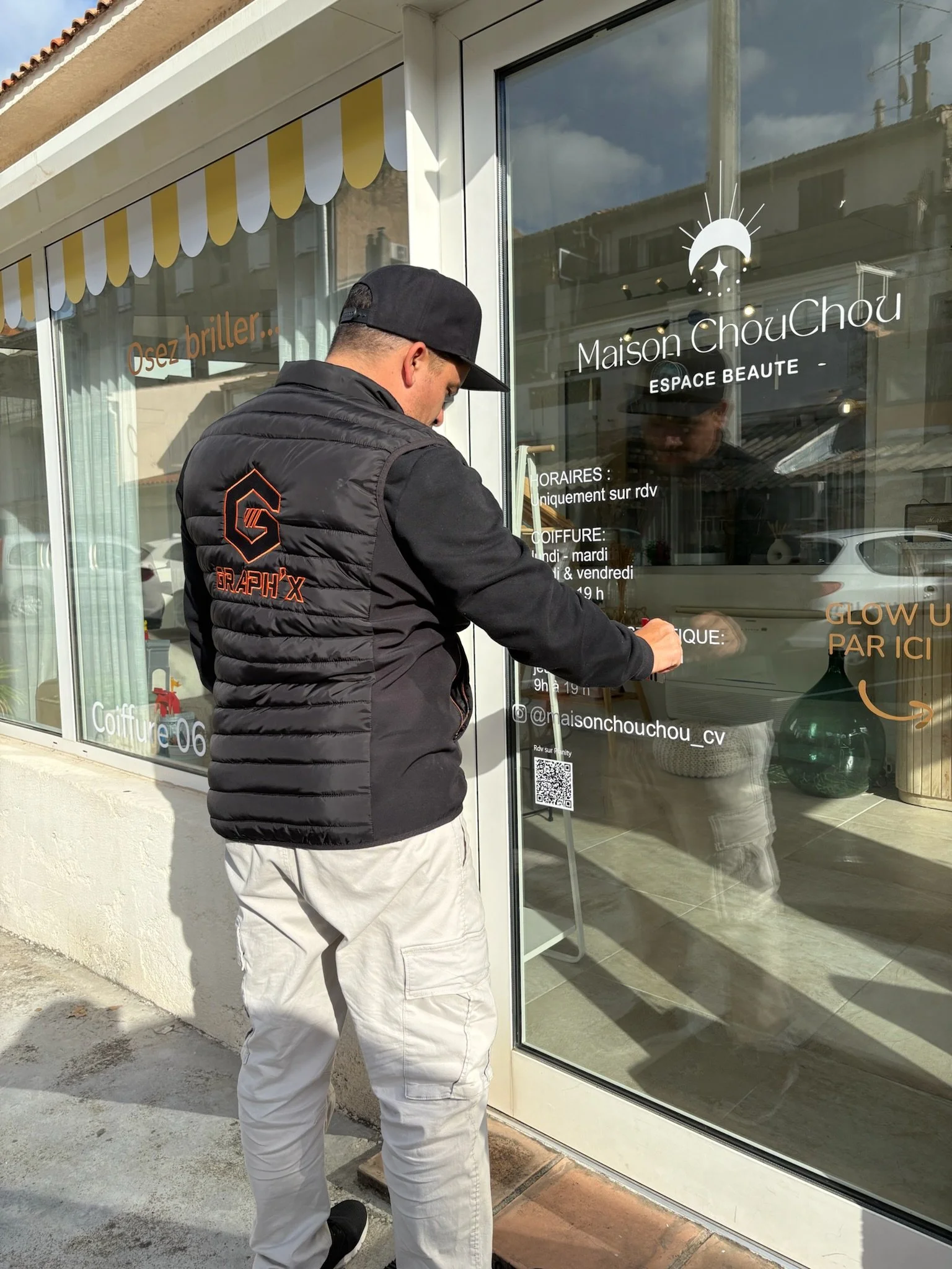 Un homme portant un sweat à capuche noir et un pantalon beige est en train de signer ou d'écrire sur une vitre avec des inscriptions en français, en face de la devanture d'un salon de beauté appelé Maison ChouChou.