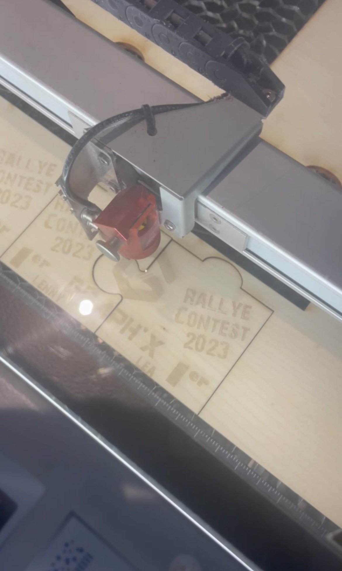 Une machine de découpe laser en train de graver le texte 'RALLYE CONTEST 2023' sur une plaque en bois ou en matière similaire.
