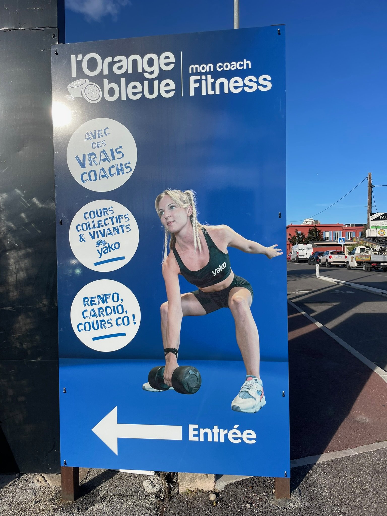 Panneau indiquant l'entrée d'une salle de fitness avec une image d'une femme soulevant un haltère. Le panneau affiche également des informations sur les cours de fitness et un slogan 'Avec des vrais coaches'.