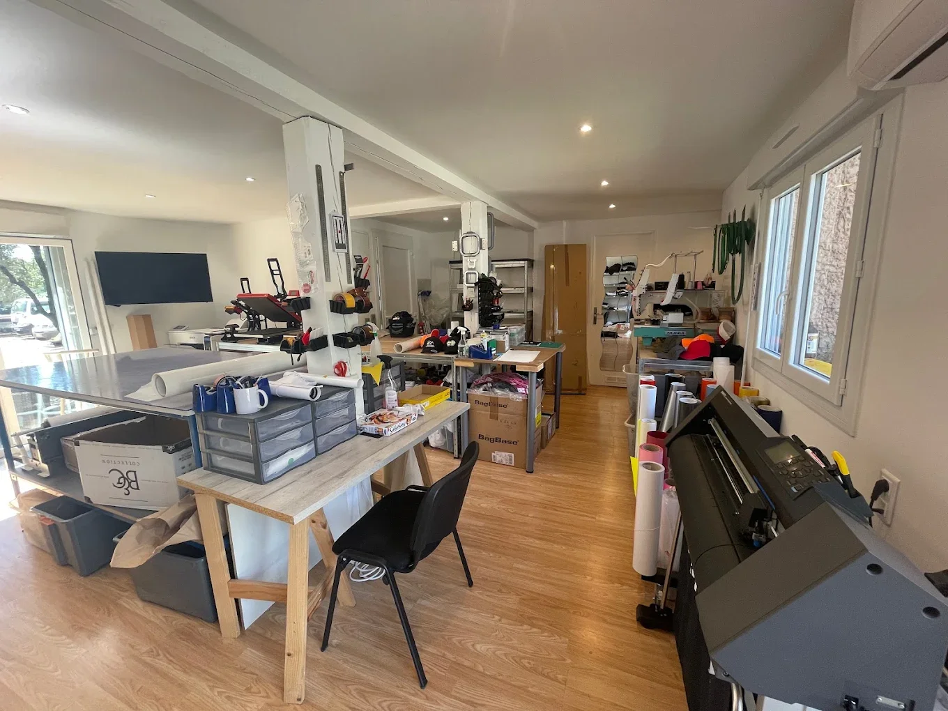Un atelier ou studio avec des tables de travail, des étagères remplies d'outils, de rouleaux de papier, des cartons, un bureau avec une chaise, un ordinateur, et une grande machine de découpe ou impression, avec des fenêtres laissant entrer la lumière.