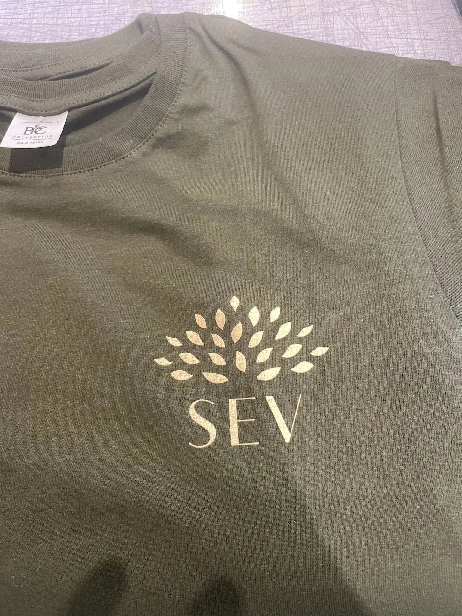 Un t-shirt gris foncé avec un logo blanc en forme de fleur stylisée et le mot 'SEV' écrit en dessous.