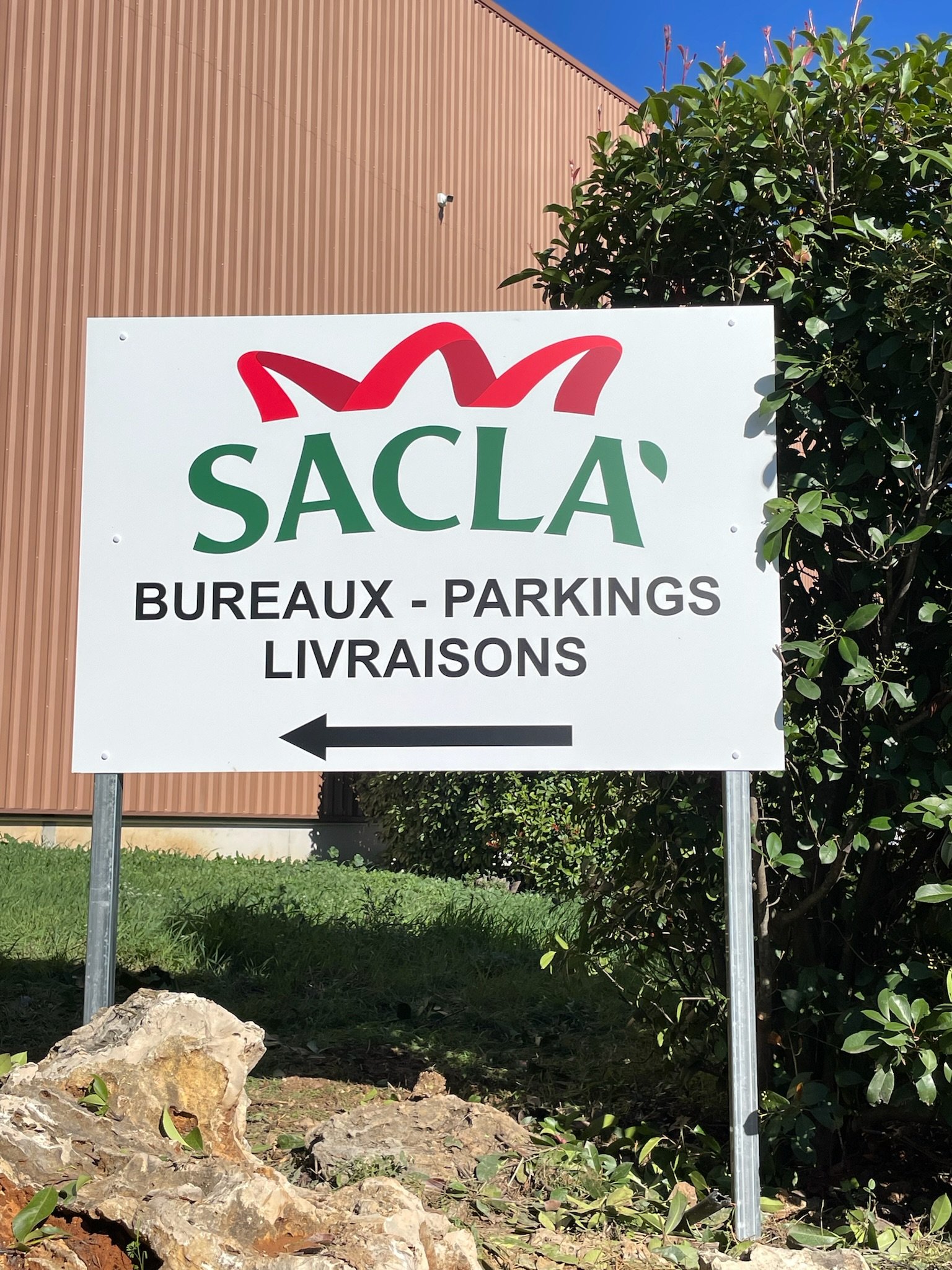 Panneau indiquant l'orientation vers les bureaux, parkings, livraisons avec une flèche pointant vers la gauche, près de buissons et de rochers.