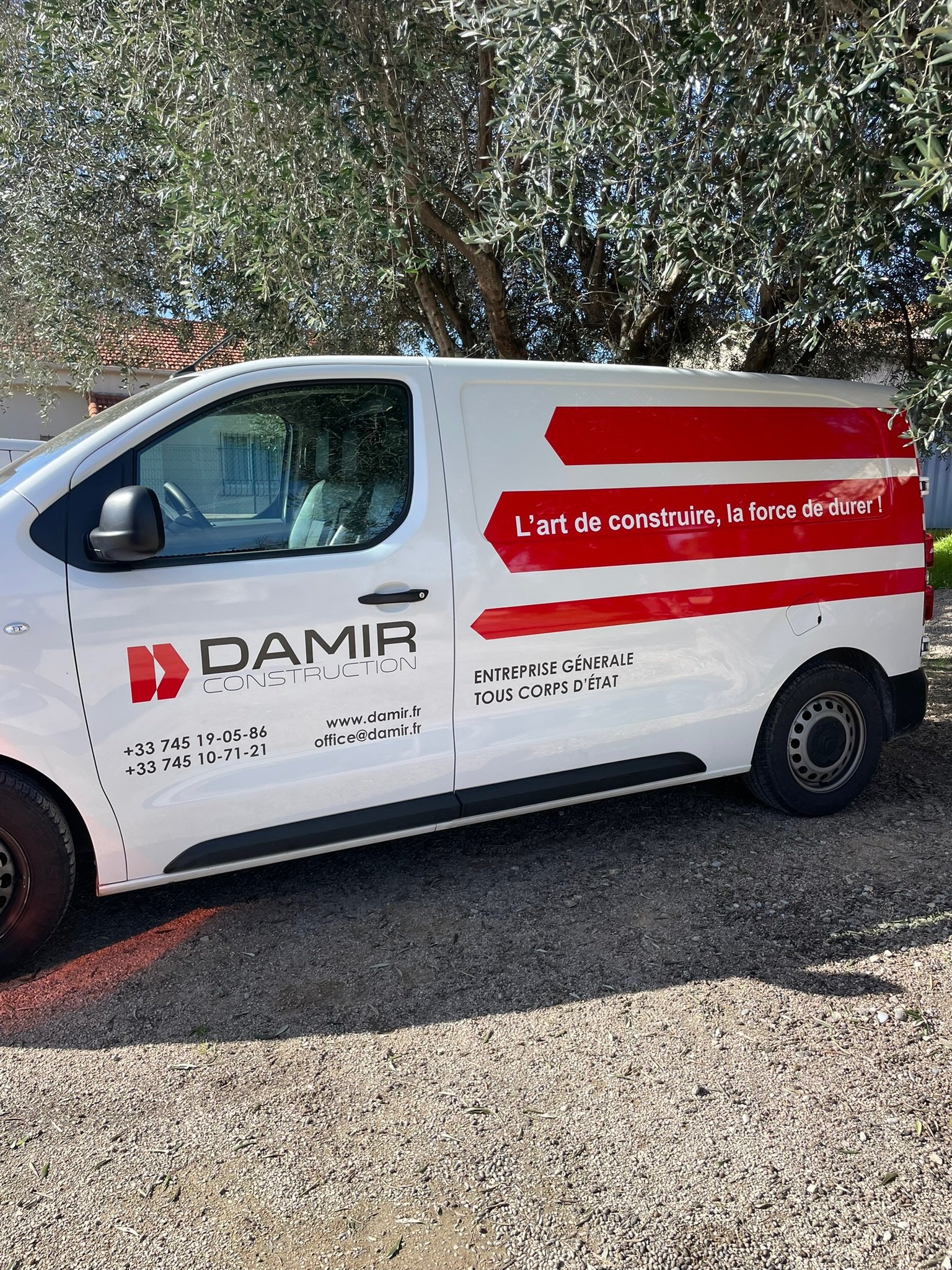 Une camionnette blanche avec des écritures et un logo rouge, stationnée sur un terrain en gravier, avec un arbre feuillu en arrière-plan.