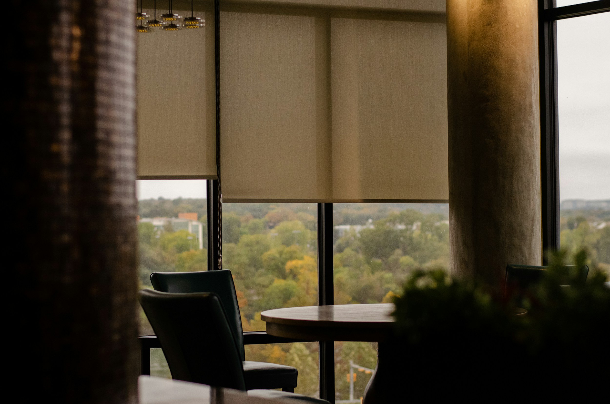 Interior of a modern office or conference room with large windows, blinds, a round wooden table, and black chairs. The window offers a view of a city with trees and buildings, suggesting an urban setting.