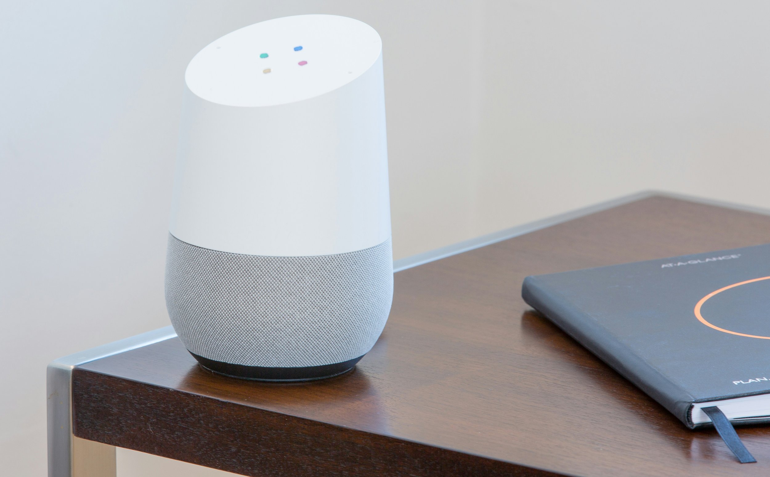 A smart speaker with a white top and gray fabric bottom placed on a wooden desk next to a black planner with an orange circular design on the cover.