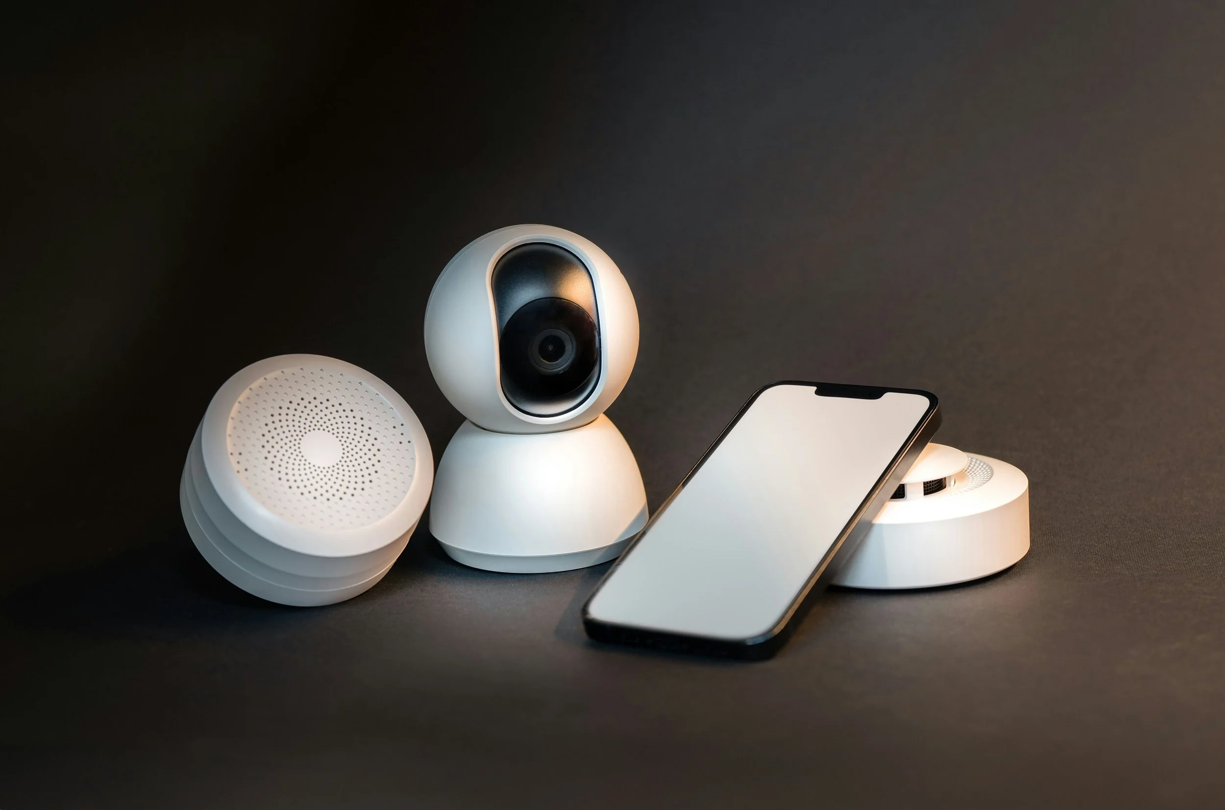 Smart home security camera, two white smart speakers, and a smartphone on a black surface.