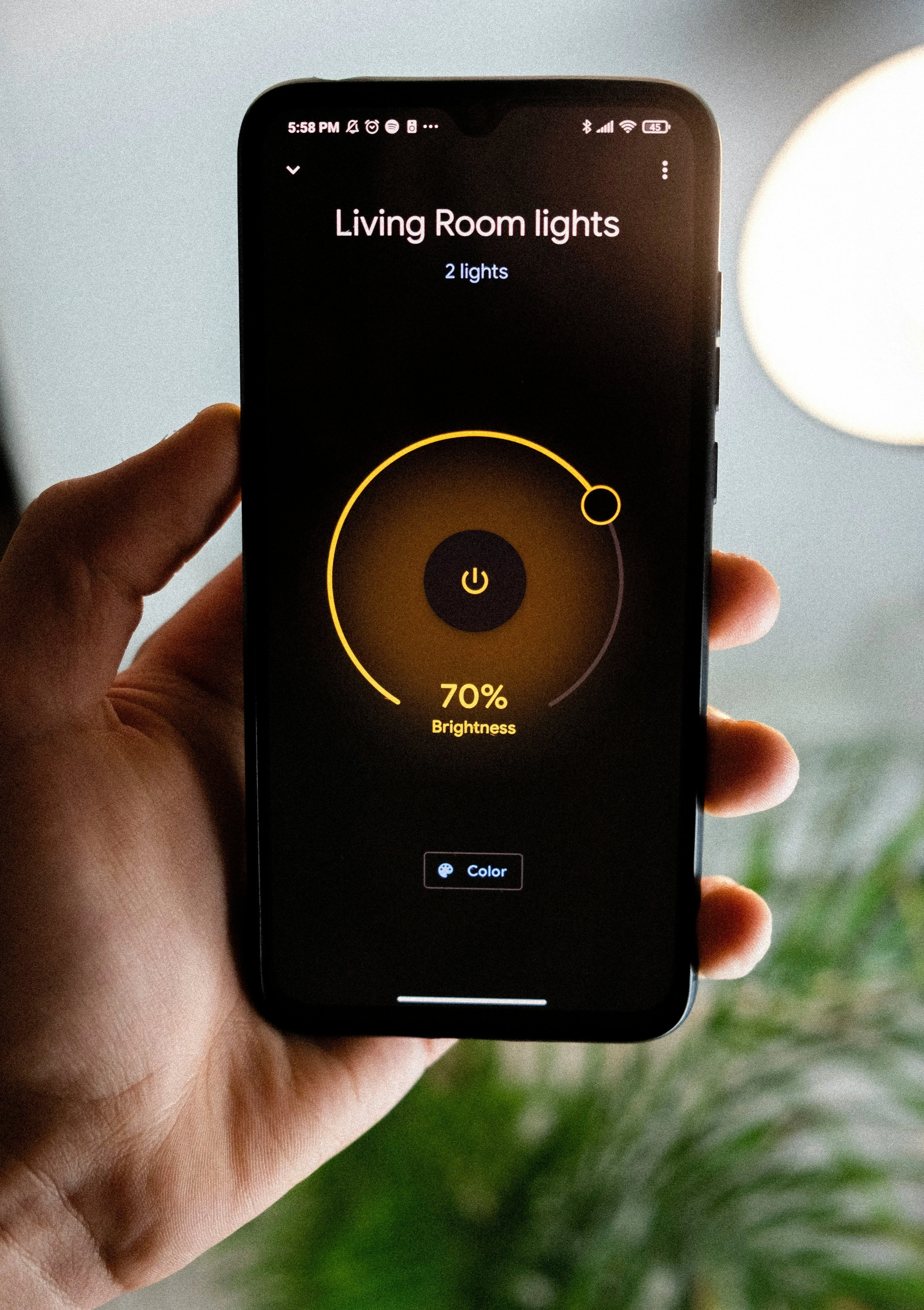 A person holding a smartphone displaying a smart home app for controlling living room lights, showing two lights at 70% brightness.