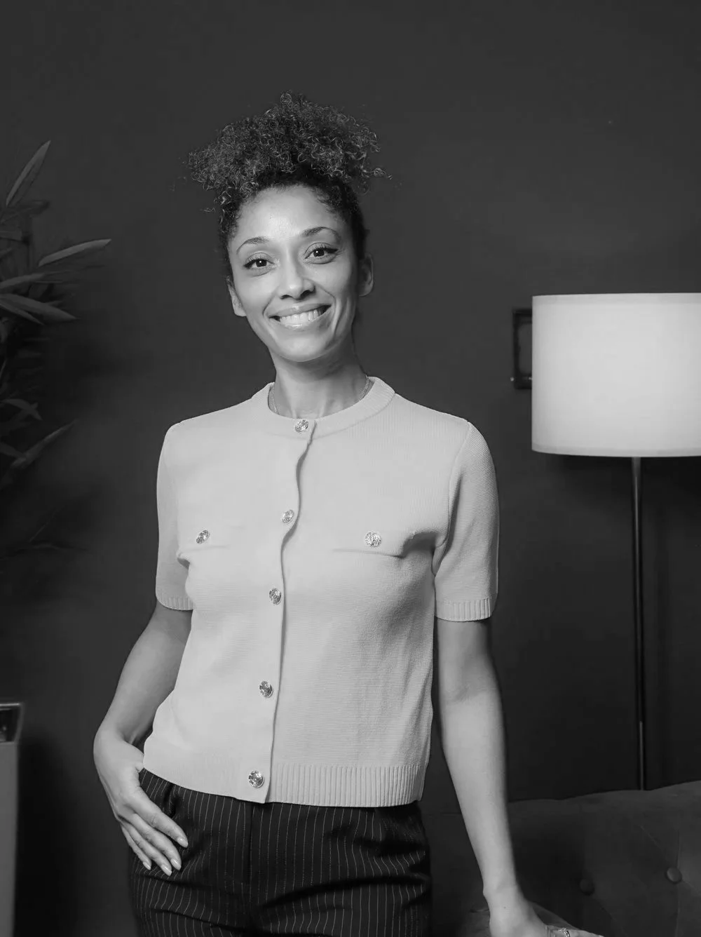 Black and white photo of a woman smiling, standing in a room with a dark wall, a plant on the left, and a floor lamp on the right.
