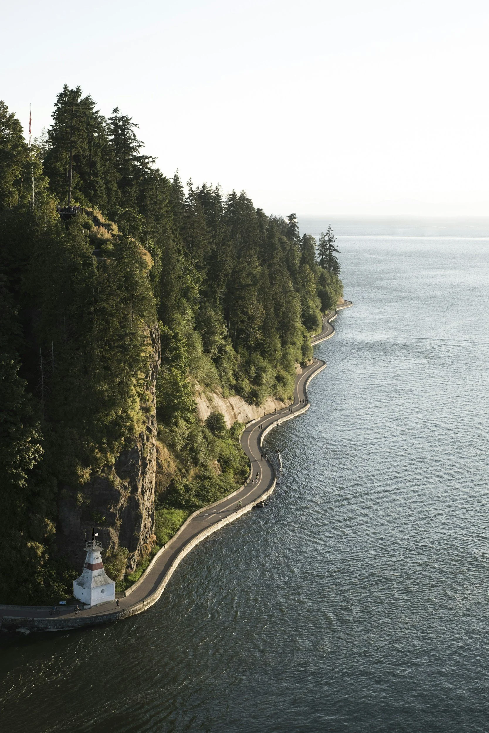 A winding road runs along a lakeshore, with a lighthouse at the bottom left and a forested hillside on the left, overlooking the water and a distant horizon.