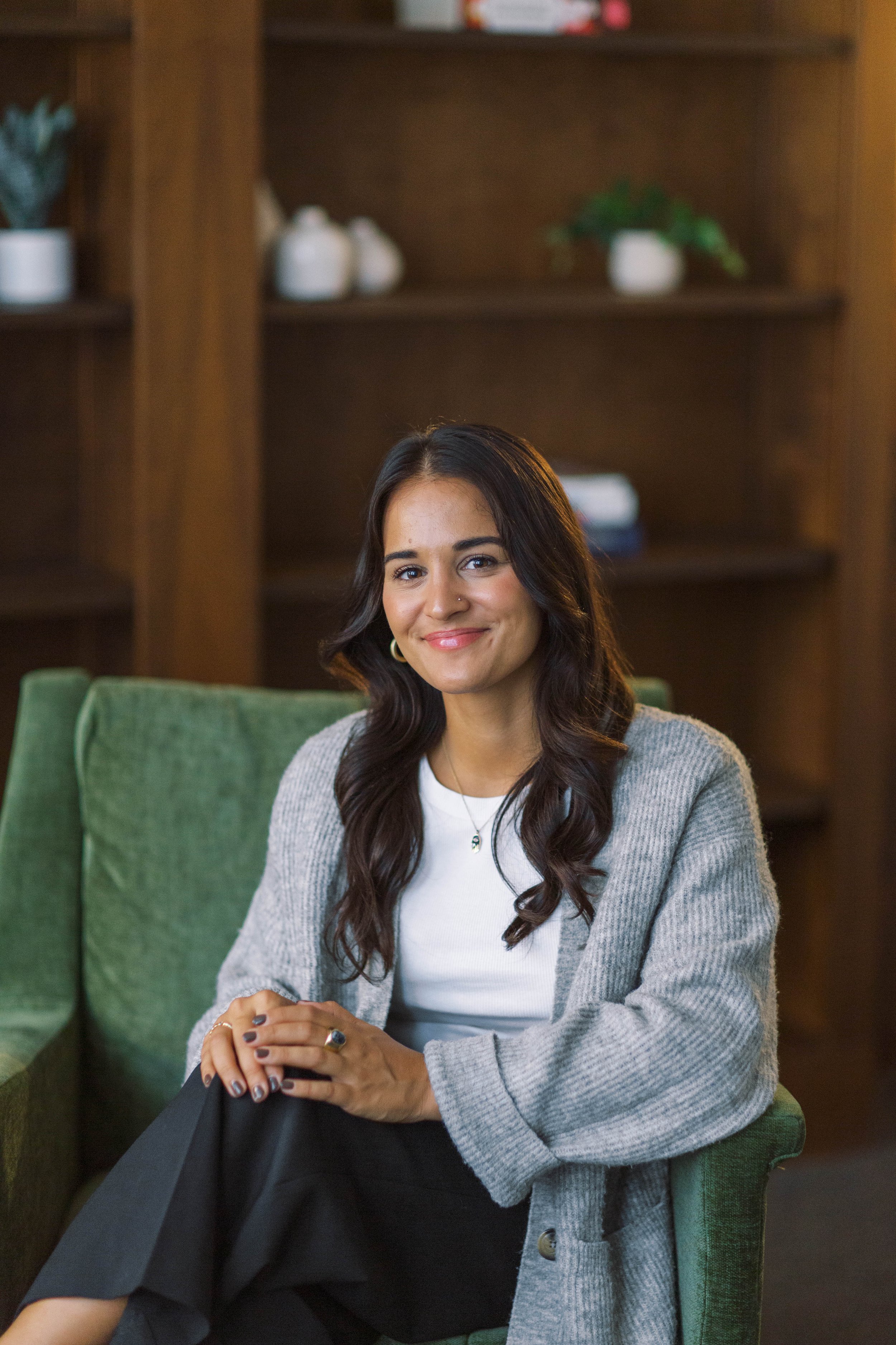 A woman with long dark hair and a nose ring sitting on a green chair in a room with wooden shelves and white decorative items.