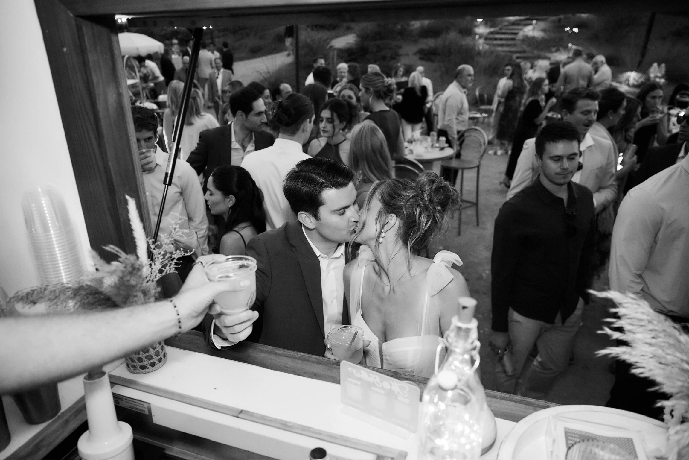 Black and white photo of a crowded party with many people socializing and dancing, seen through a window or mirror. Two people are kissing in the foreground, holding drinks. The scene appears lively and celebratory.