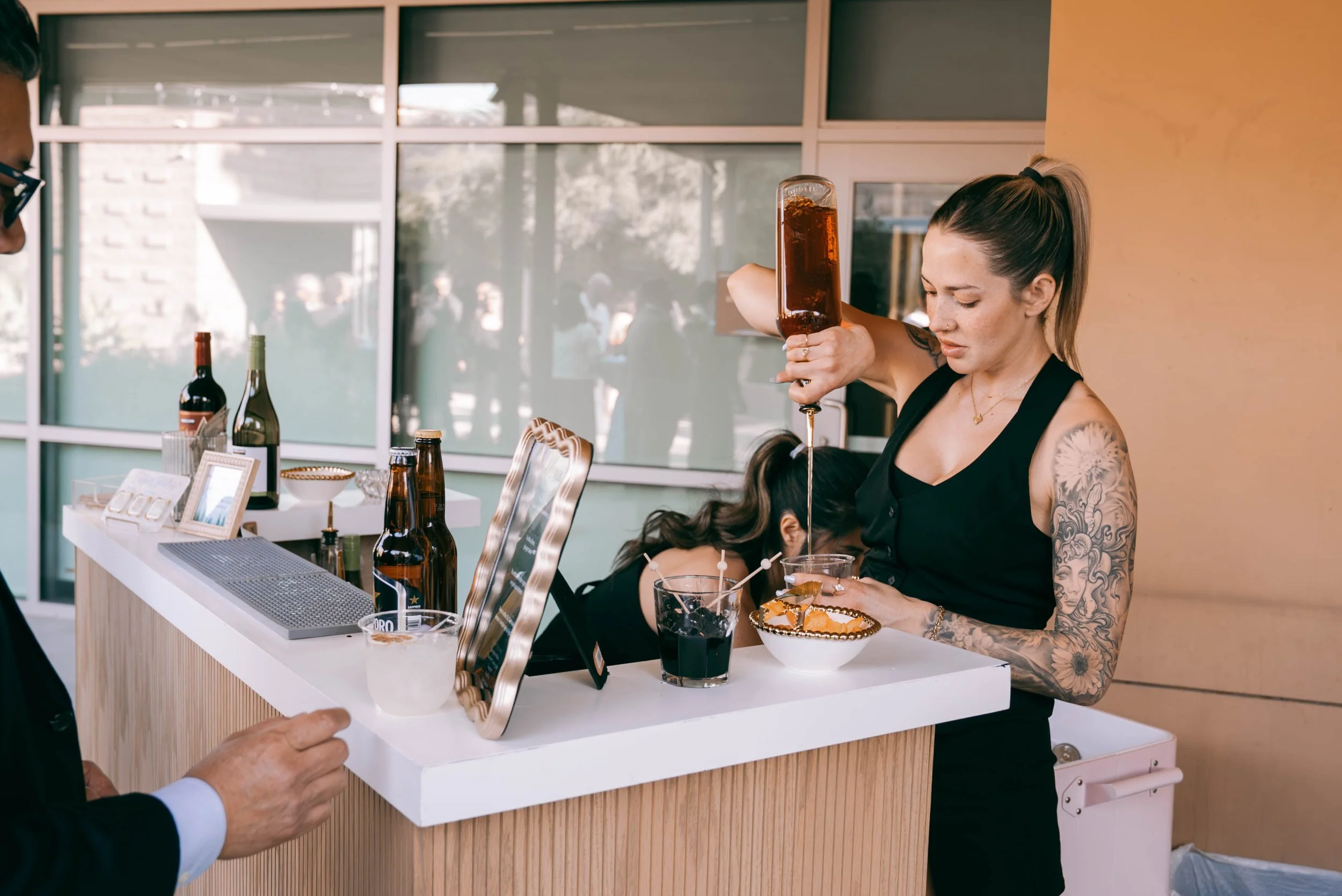 A woman with tattoos on her arm making a cocktail at a bar, pouring a dark liquid from a large bottle into a glass with a garnish, while another person is partially visible in the foreground. The bar has bottles, a framed photo, and a mirror behind i