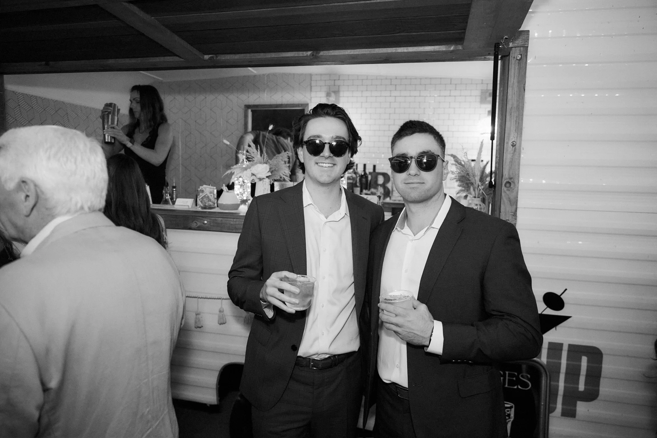 Two men in suits wearing sunglasses, holding drinks, at a party or celebration. People and decorations visible in the background.