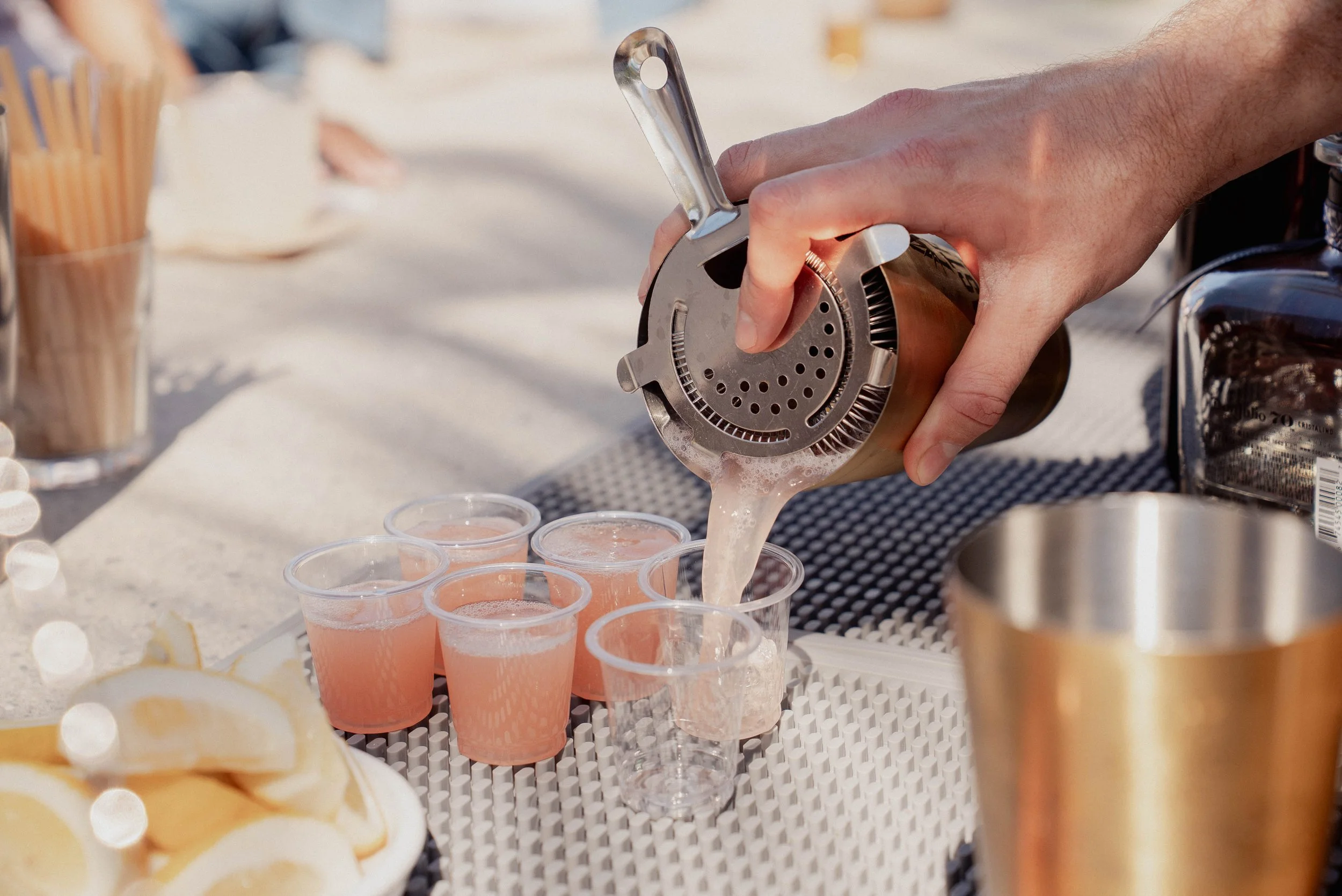 Person pouring grapefruit juice into small glasses at an outdoor gathering with lemon slices and utensils in the background.
