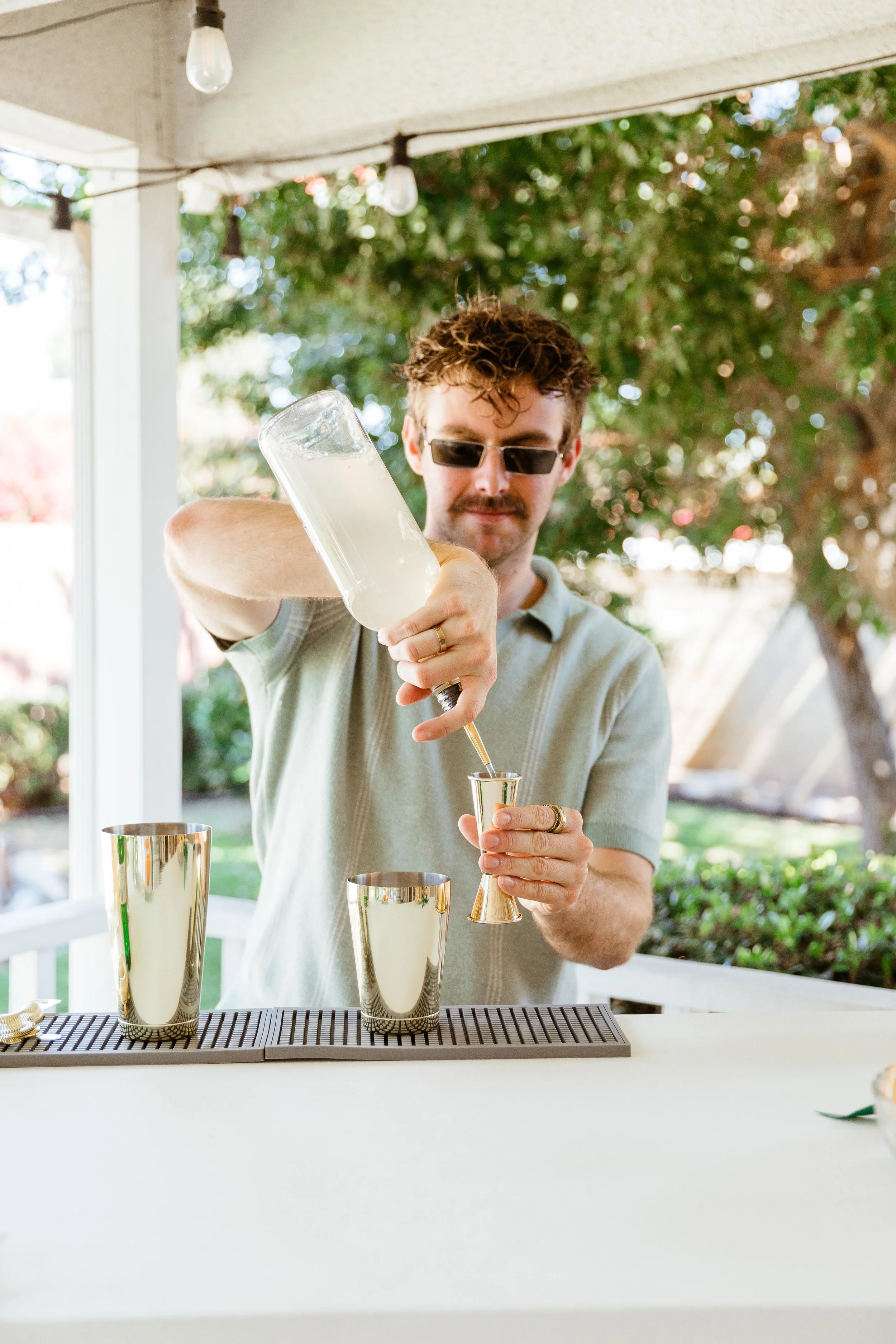 Man with sunglasses pouring drinks into a shot glass at an outdoor bar.