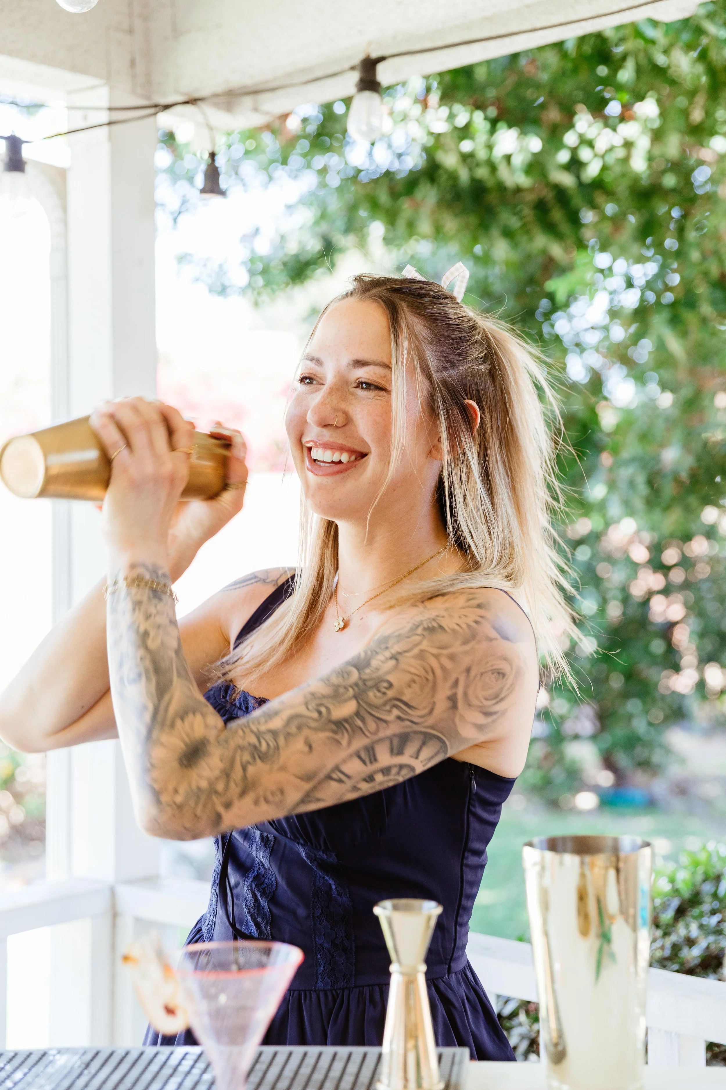 A smiling woman with tattoos on her arms, holding a cocktail shaker, preparing drinks outdoors.