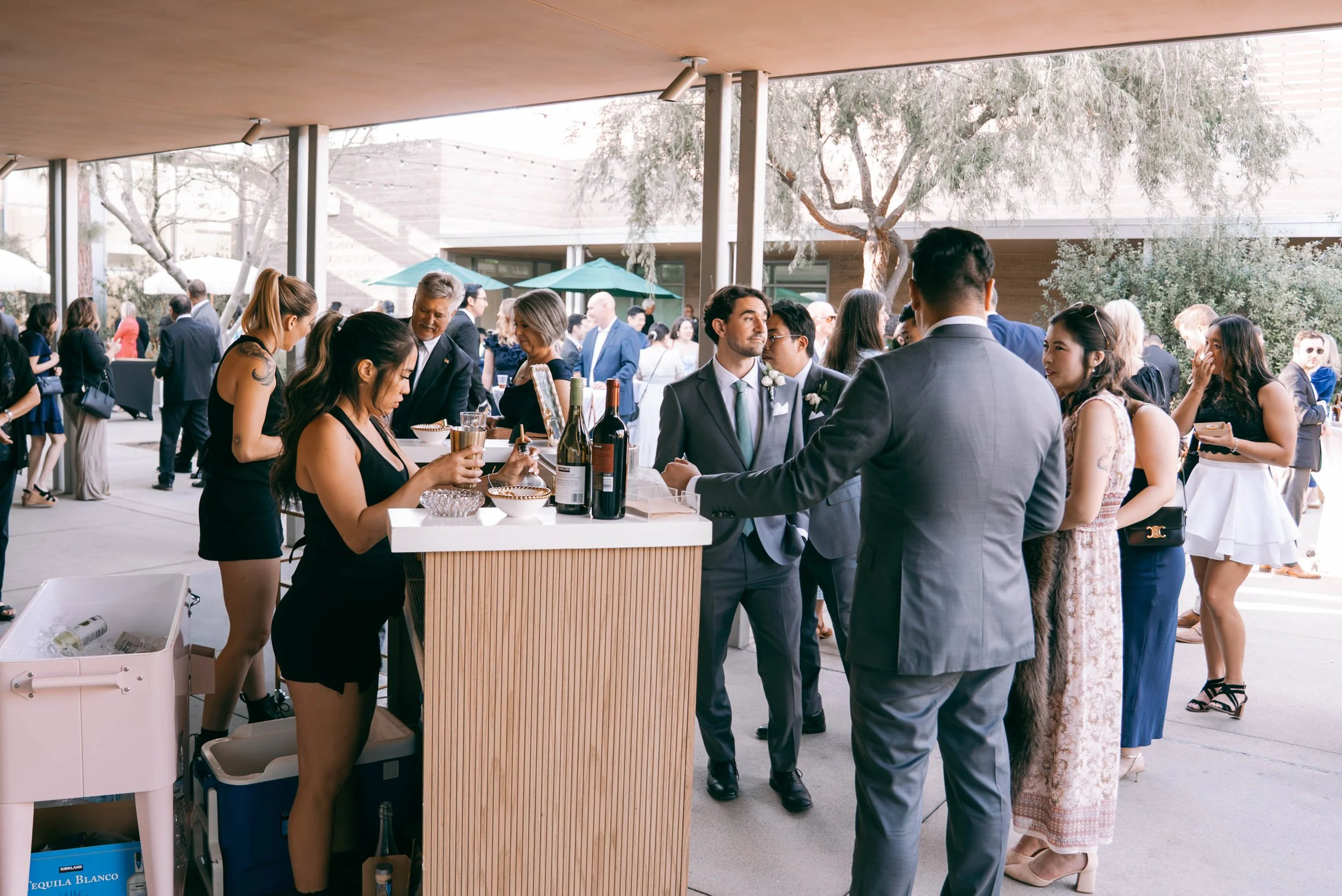 People dressed in formal attire socializing at an outdoor wedding reception, with a bar and drinks in the foreground and trees in the background.