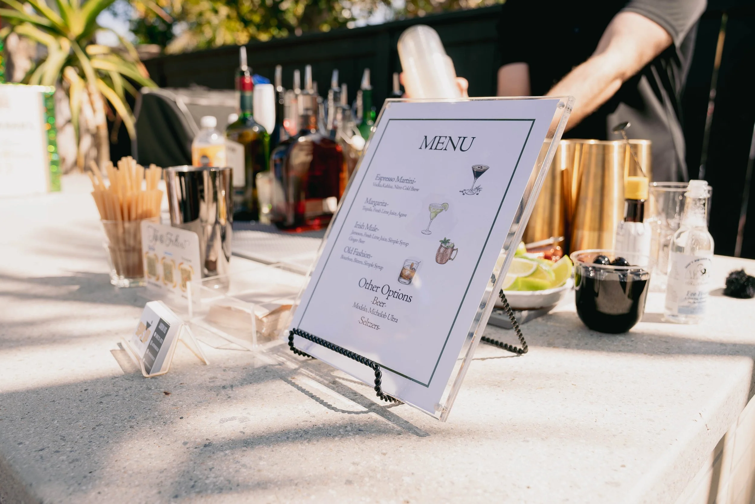Bar setup with a menu board, liquor bottles, cocktail ingredients, and garnishes on a outdoor countertop