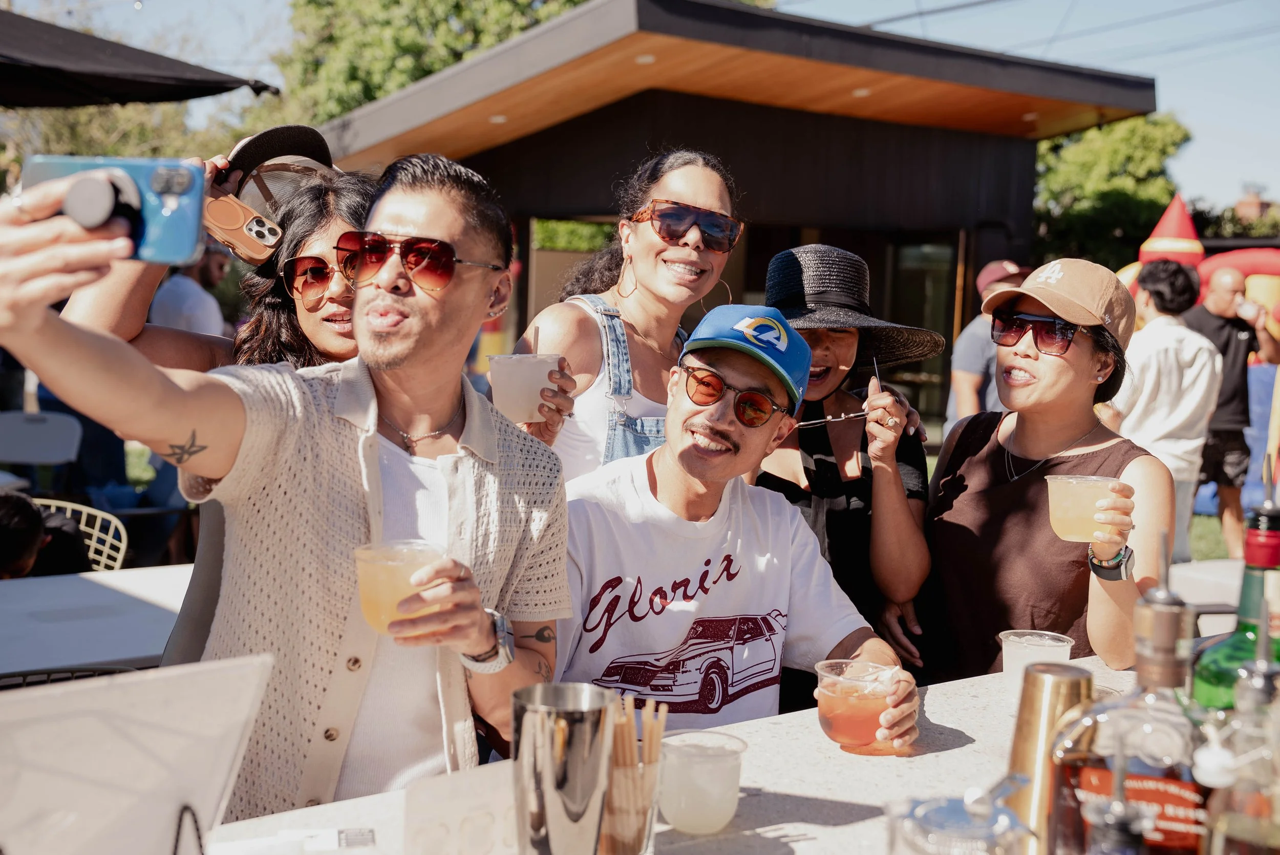 A group of friends taking a selfie at an outdoor gathering on a sunny day. They are smiling, wearing sunglasses, and holding drinks. The background shows more people and a modern house with a green yard.