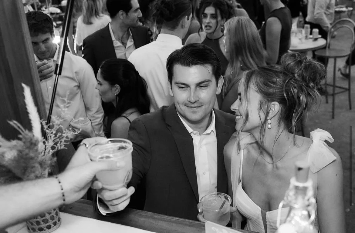 A black and white photograph of a social gathering or party with many people in the background. In the foreground, a man in a suit is smiling and holding a drink, while a woman with curly hair and earrings is leaning in, smiling at him. Someone's han