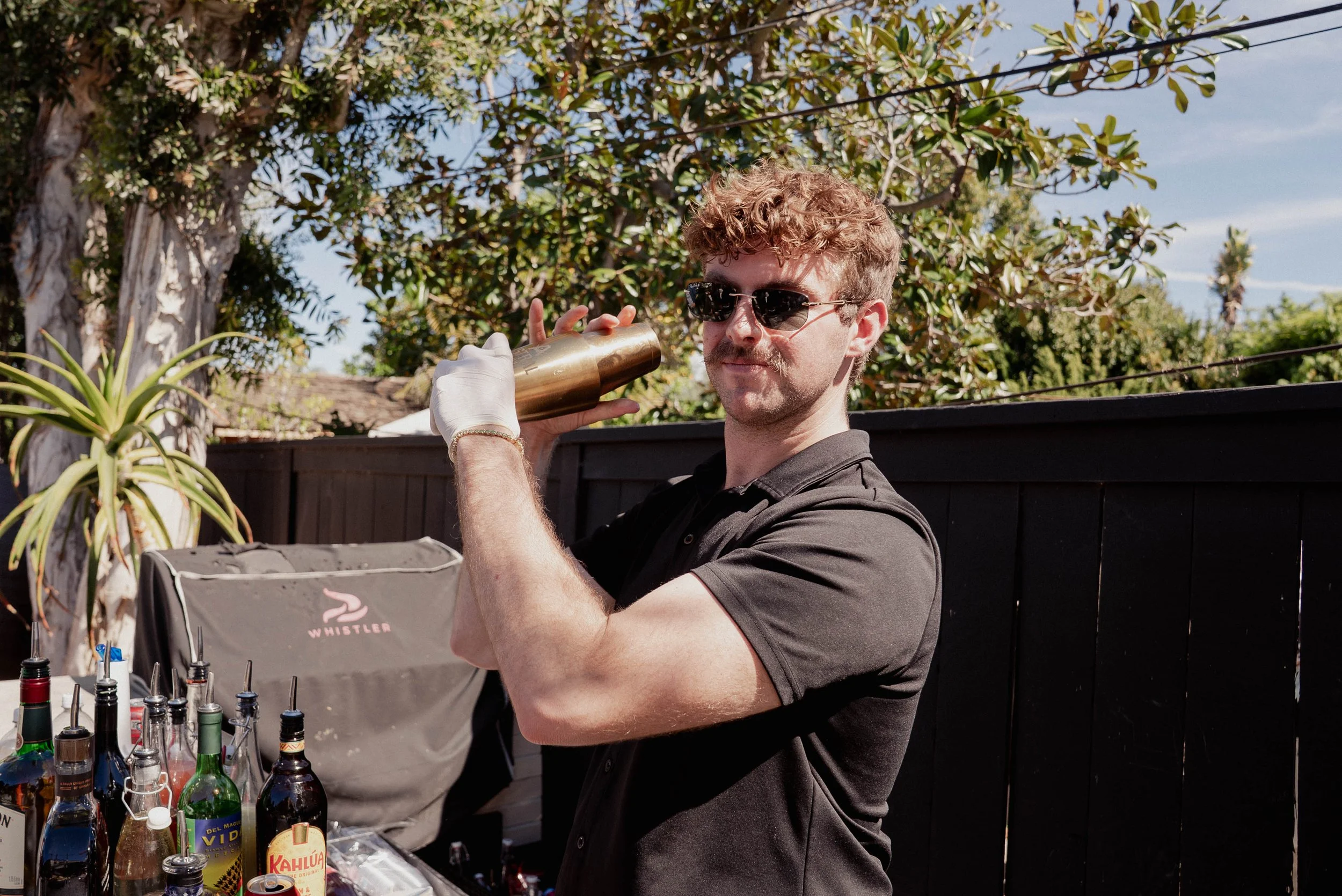 A man with curly hair, wearing sunglasses and a black shirt, holding a gold cocktail shaker on his shoulder outdoors next to a bar setup, with bottles and a black fence in the background.
