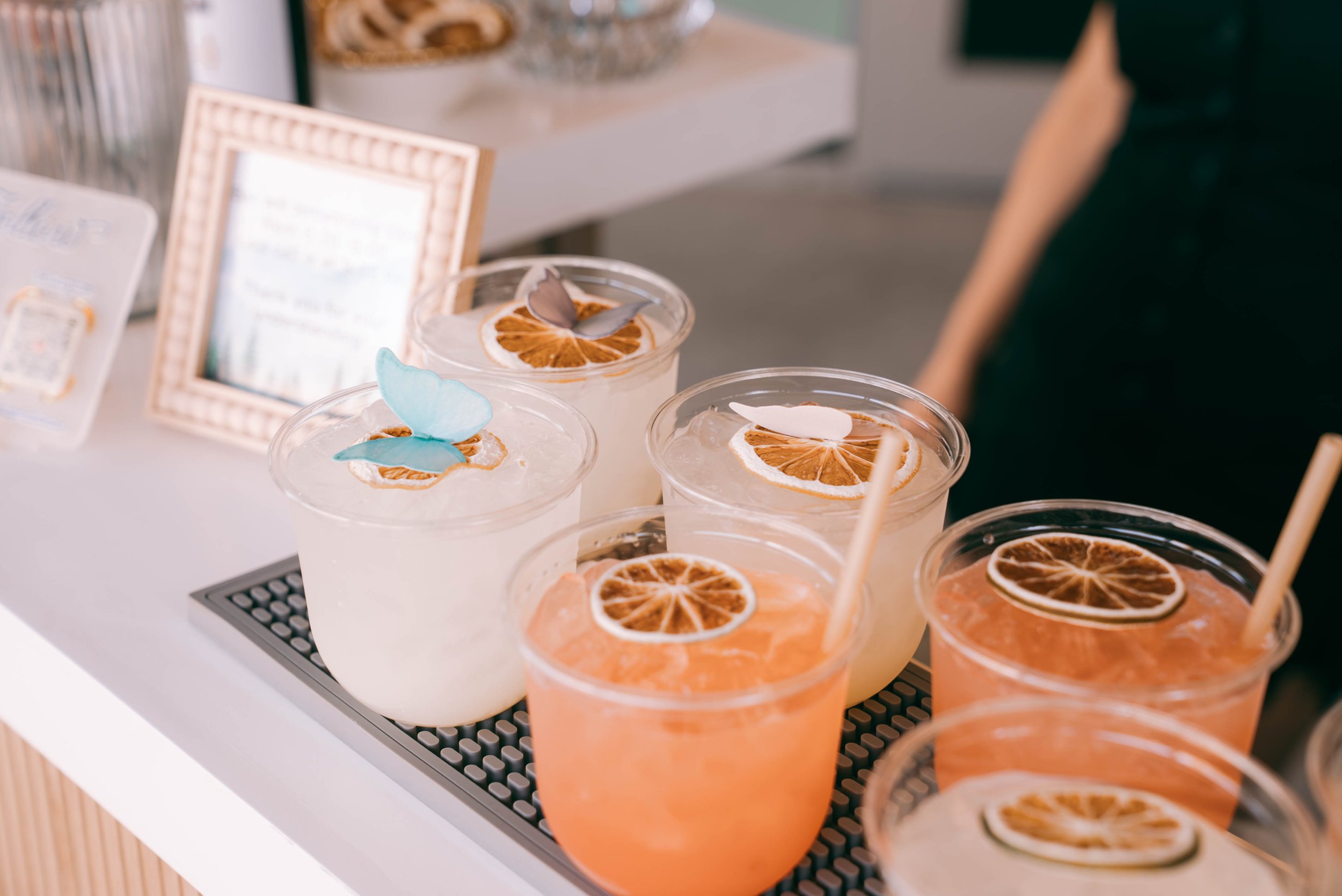 Multiple glasses of citrus-flavored beverages with dried orange slices and decorative lemon and butterfly paper cutouts, displayed on a counter at a drink stand.