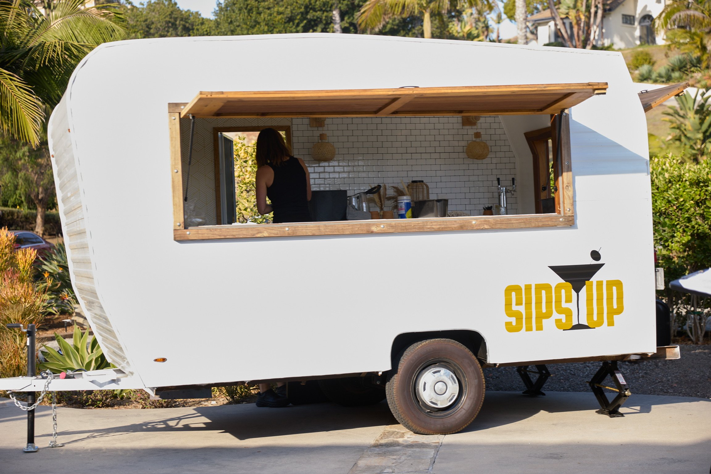 A white mobile bar trailer with a yellow logo reading 'SIPS UP' and a martini glass icon, parked outdoors with a woman inside preparing drinks.