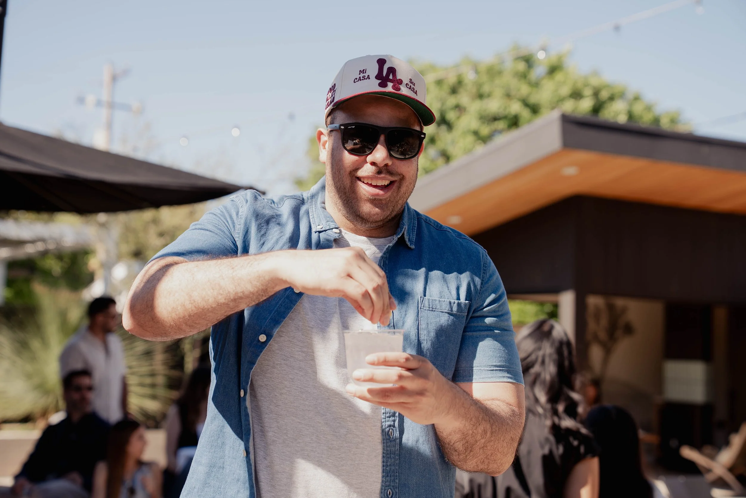 Man smiling in sunglasses, wearing a baseball cap and denim shirt, pouring a drink at an outdoor gathering.