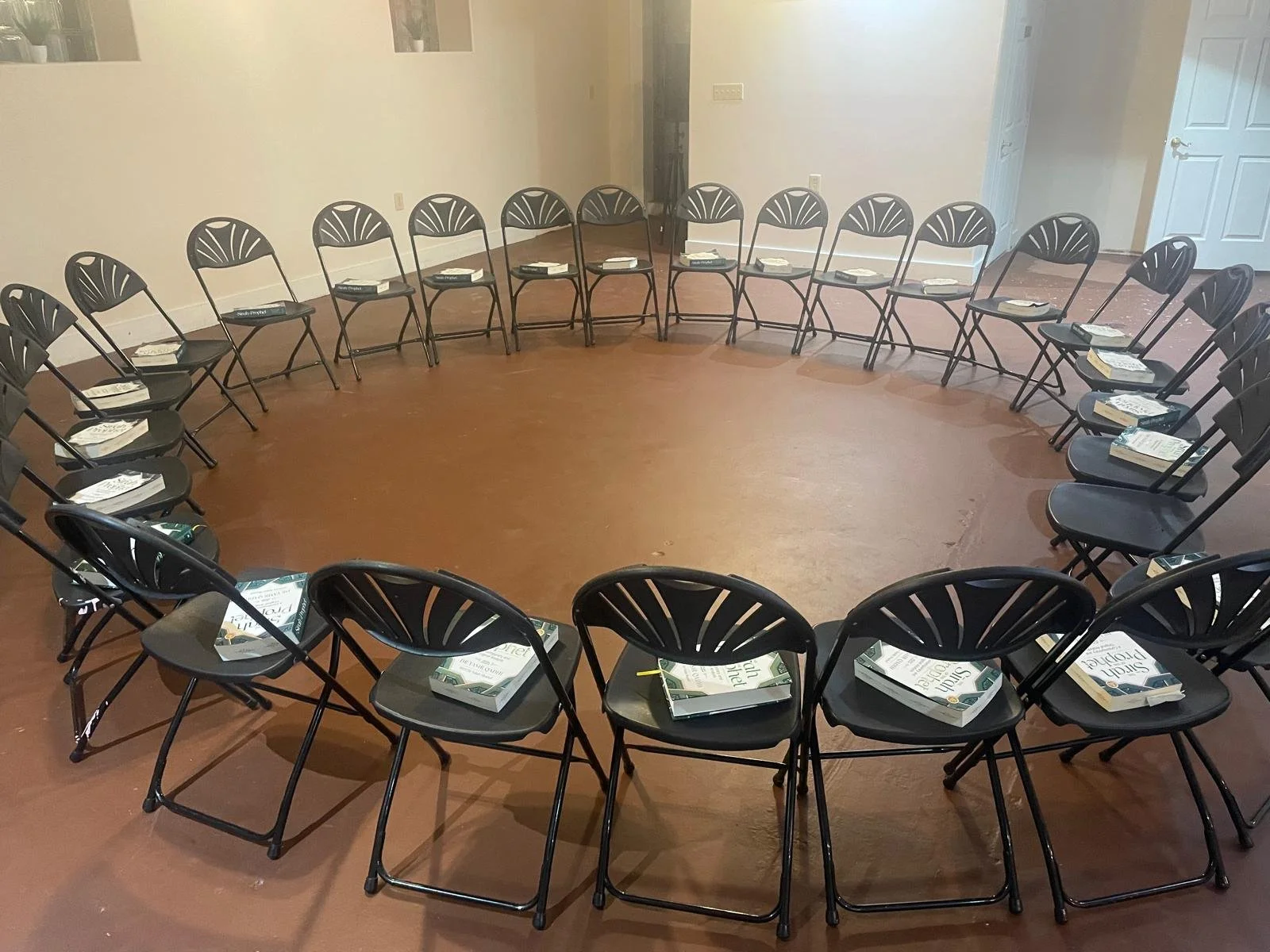 A circular arrangement of black chairs with paper and booklets on each seat in a room with wooden floor and plain white walls.