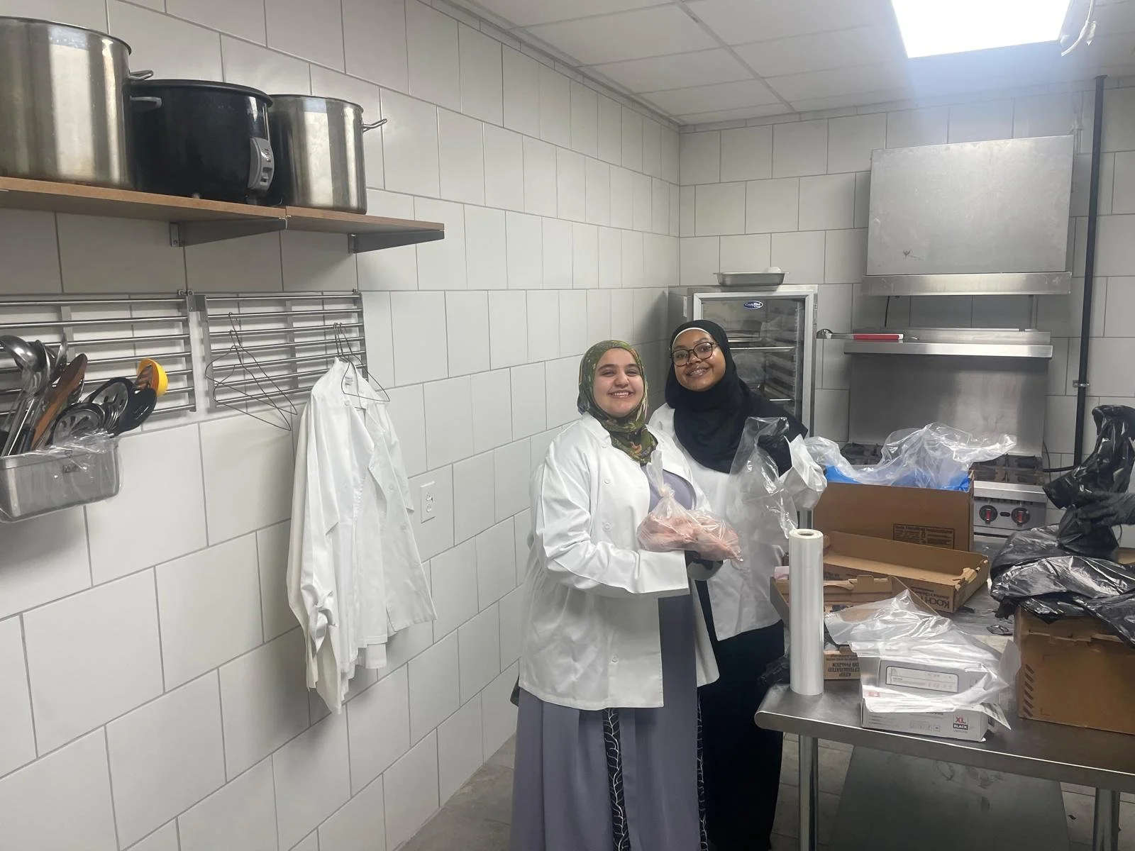 Two women wearing white chef coats and hijabs smiling in a commercial kitchen surrounded by packaging materials and kitchen equipment.