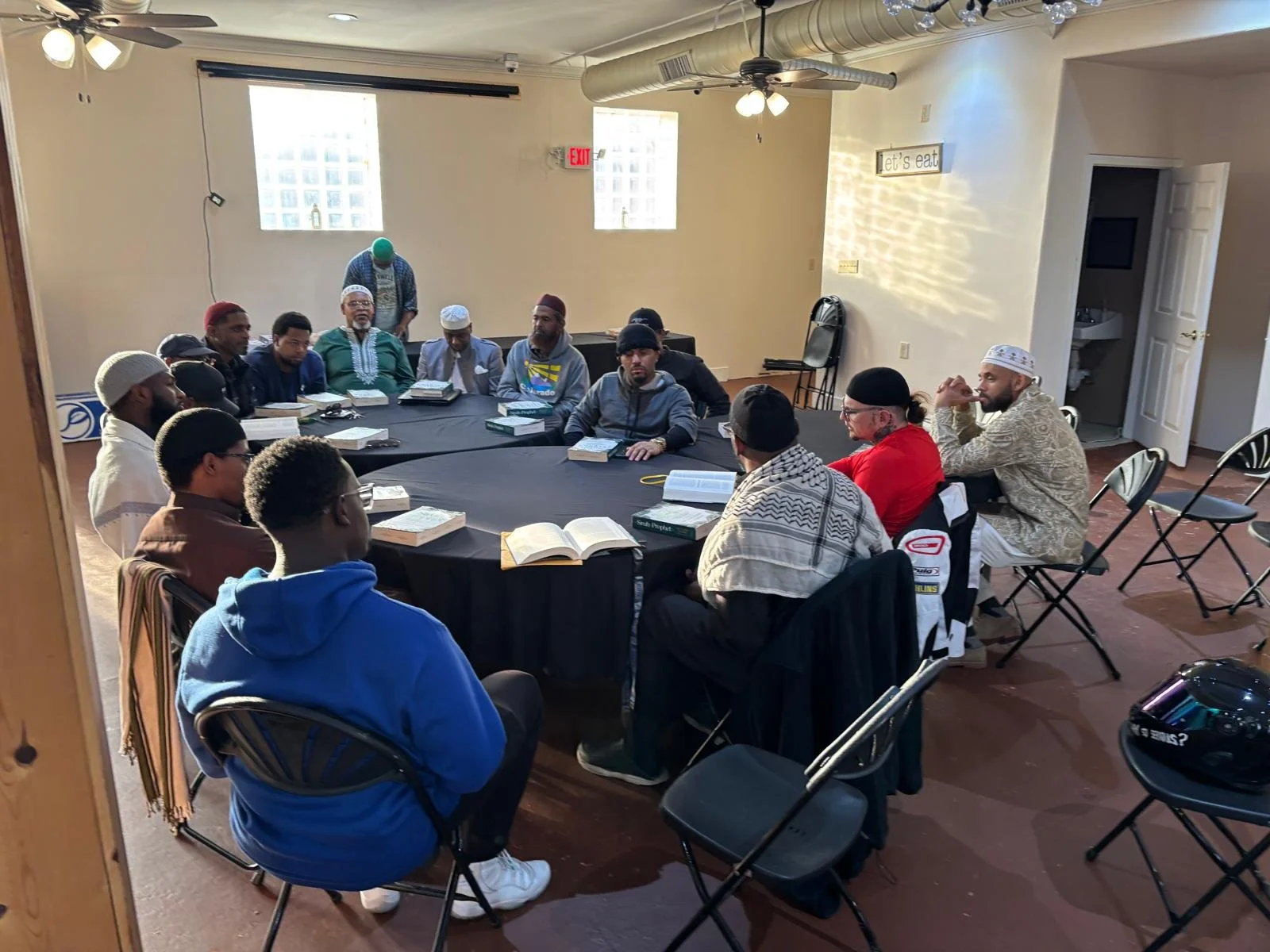 A diverse group of men sitting around a large round table engaged in discussion, with books and notebooks in front of them, in a well-lit indoor space.