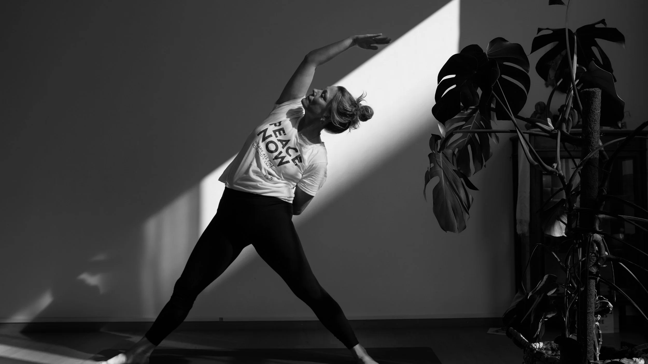 A woman practicing yoga indoors, stretching with her left arm overhead and body arched back, in front of a plain wall with a large plant and bicycle to the right.