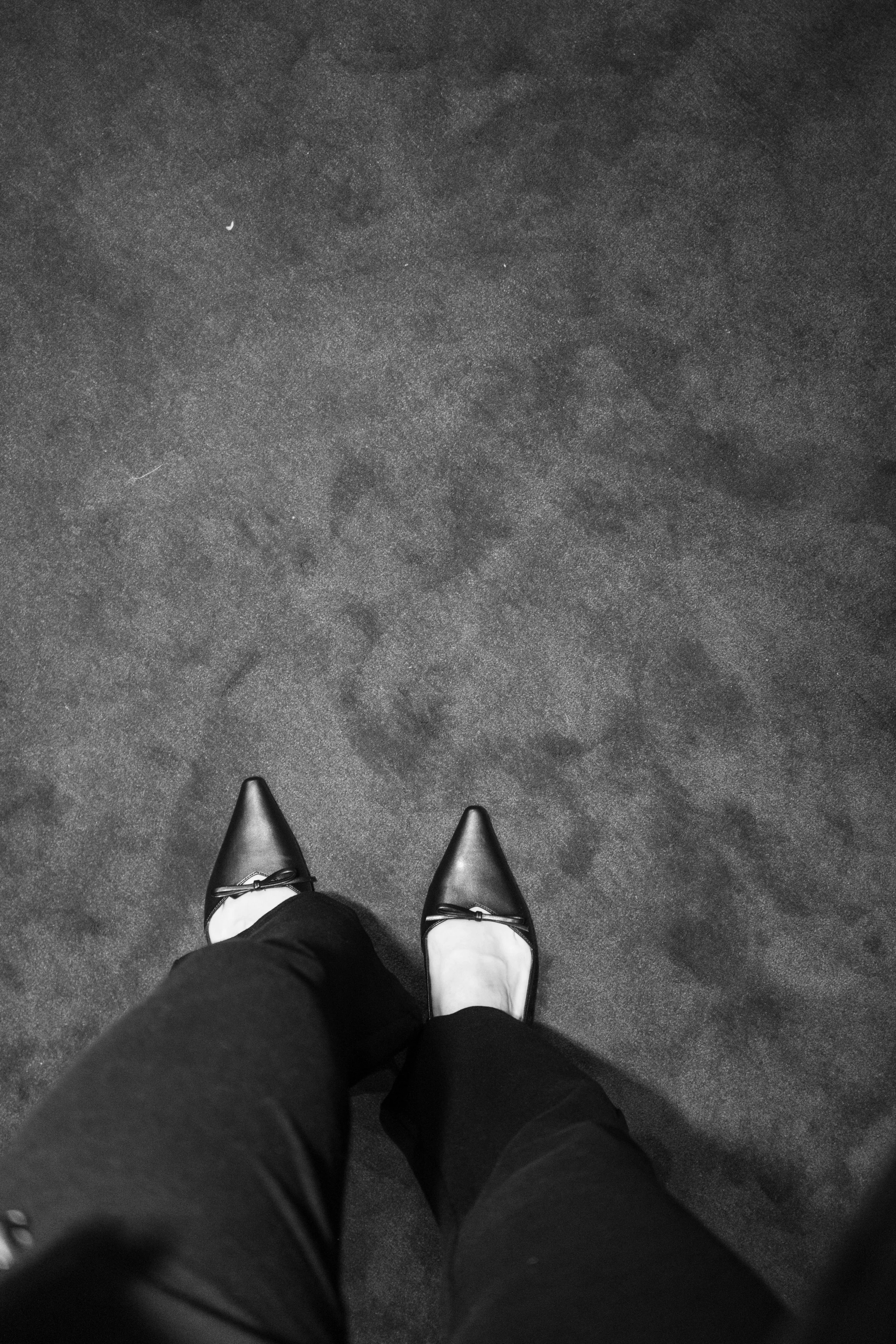 View of a person standing on a textured dark gray carpet, wearing black pointed-toe shoes with small bows and light-colored socks, with a portion of black pants visible.