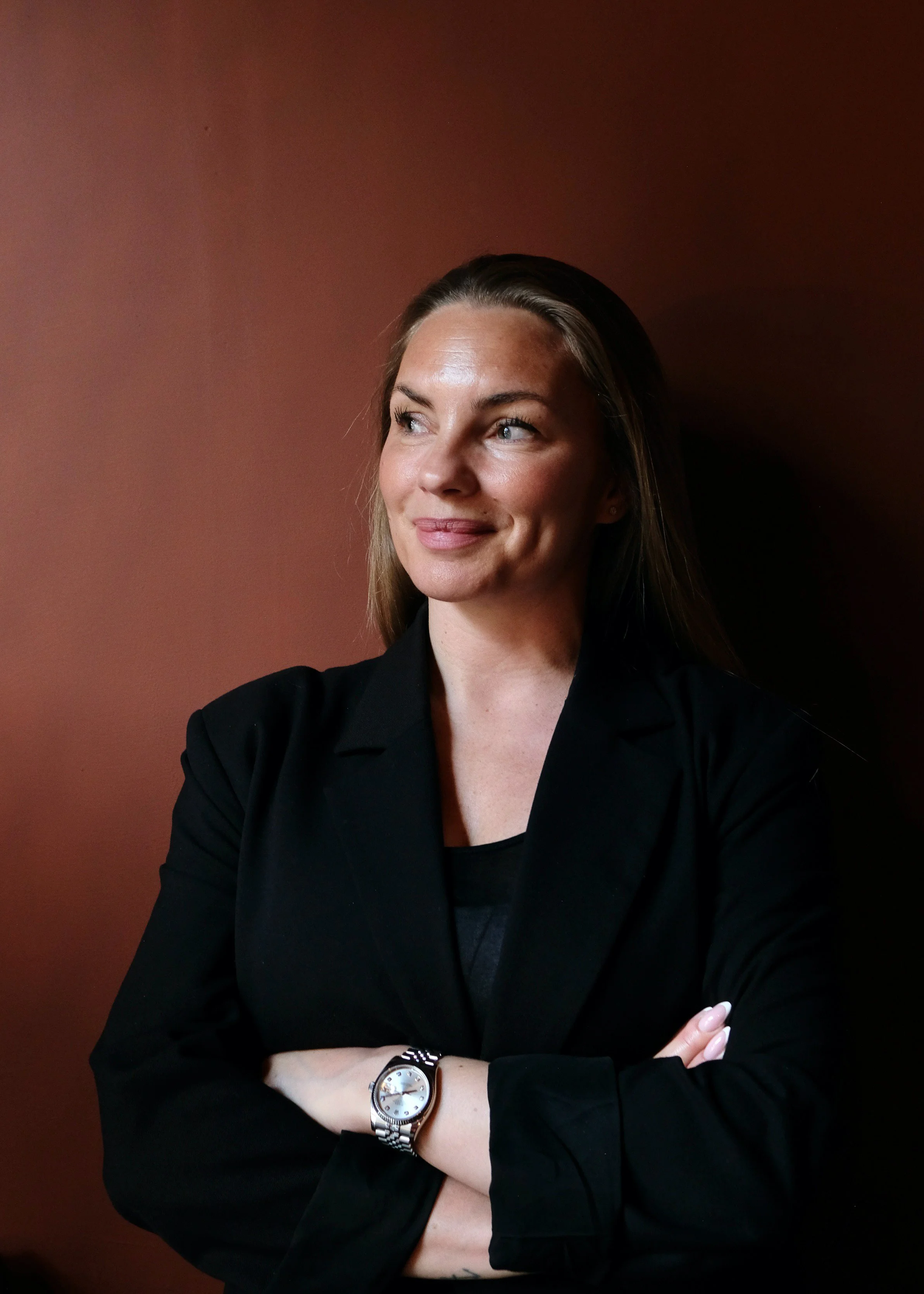 A woman with brown hair and blue eyes wearing a black blazer and a silver watch, standing against a plain reddish-brown wall, looking to her left with crossed arms.