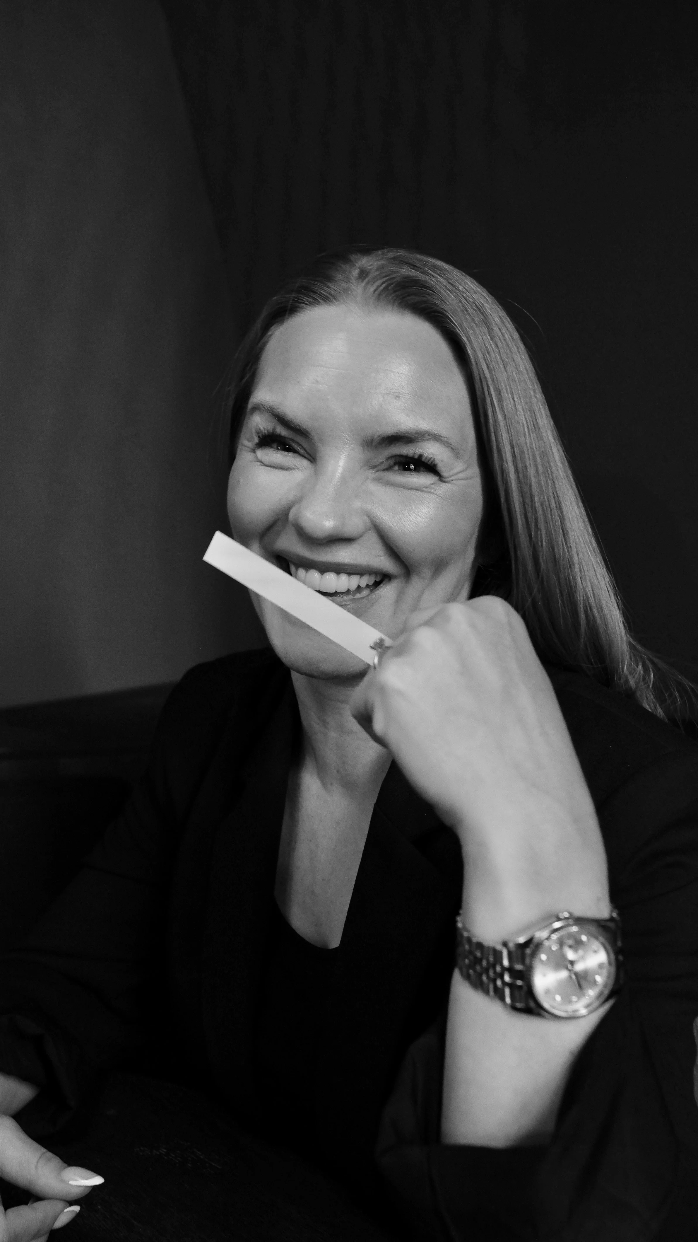 A black and white photo of a smiling woman with long hair, wearing a watch, holding a piece of paper near her mouth, sitting at a table in a dark room.