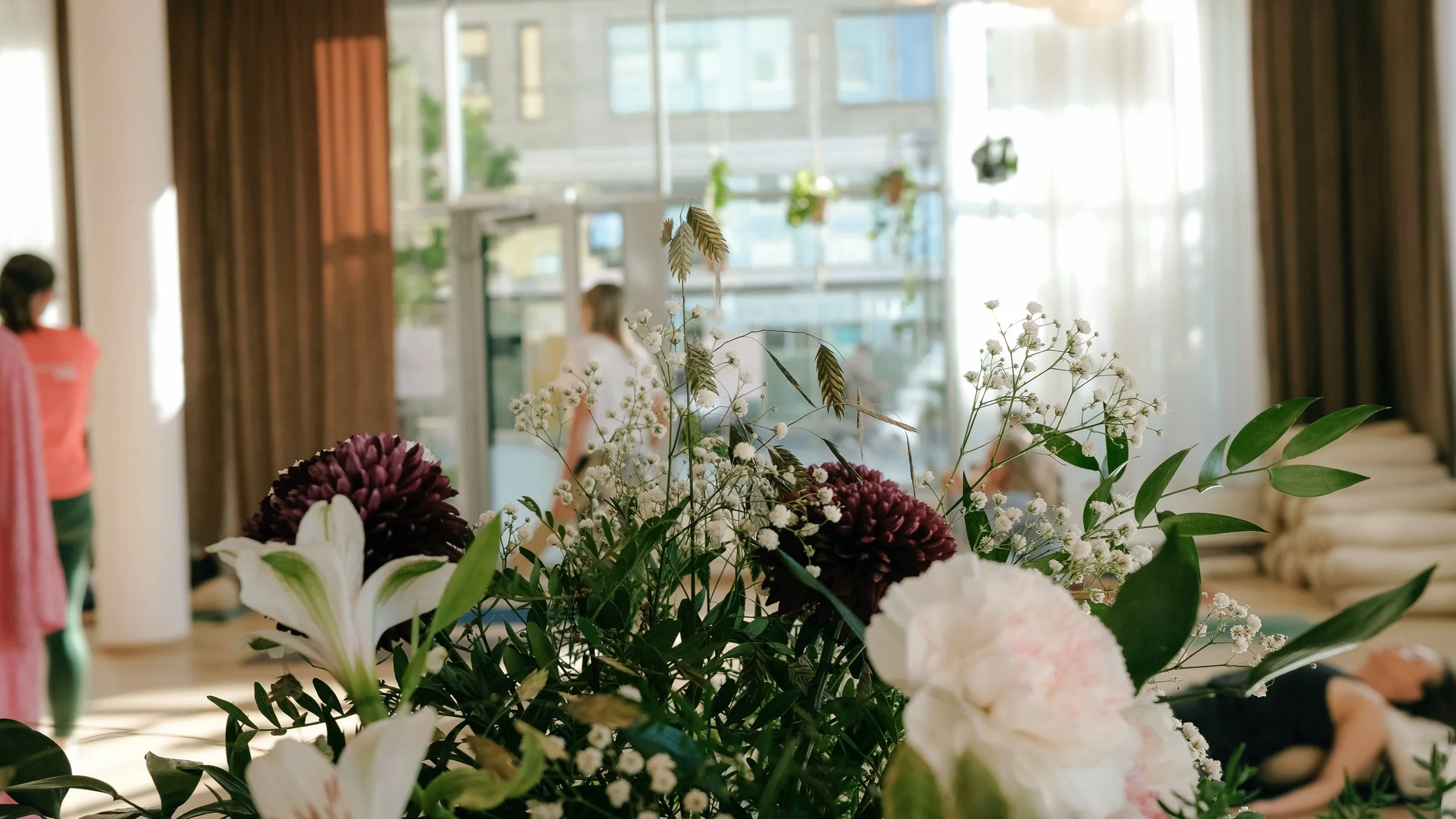 A close-up of an arrangement of flowers, including white lilies, pink roses, purple dahlias, and small white filler flowers, in an indoor setting with large windows and curtains in the background.