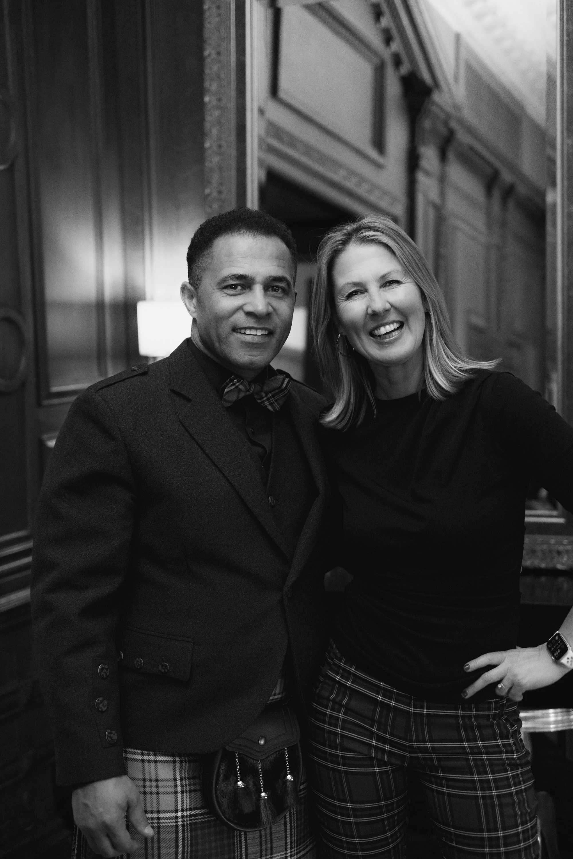 Black and white photo of two people smiling and posing together in an indoor setting with wooden wall panels and framed artwork in the background. The person on the left is wearing a dark blazer and a plaid skirt, with a small purse hanging from thei
