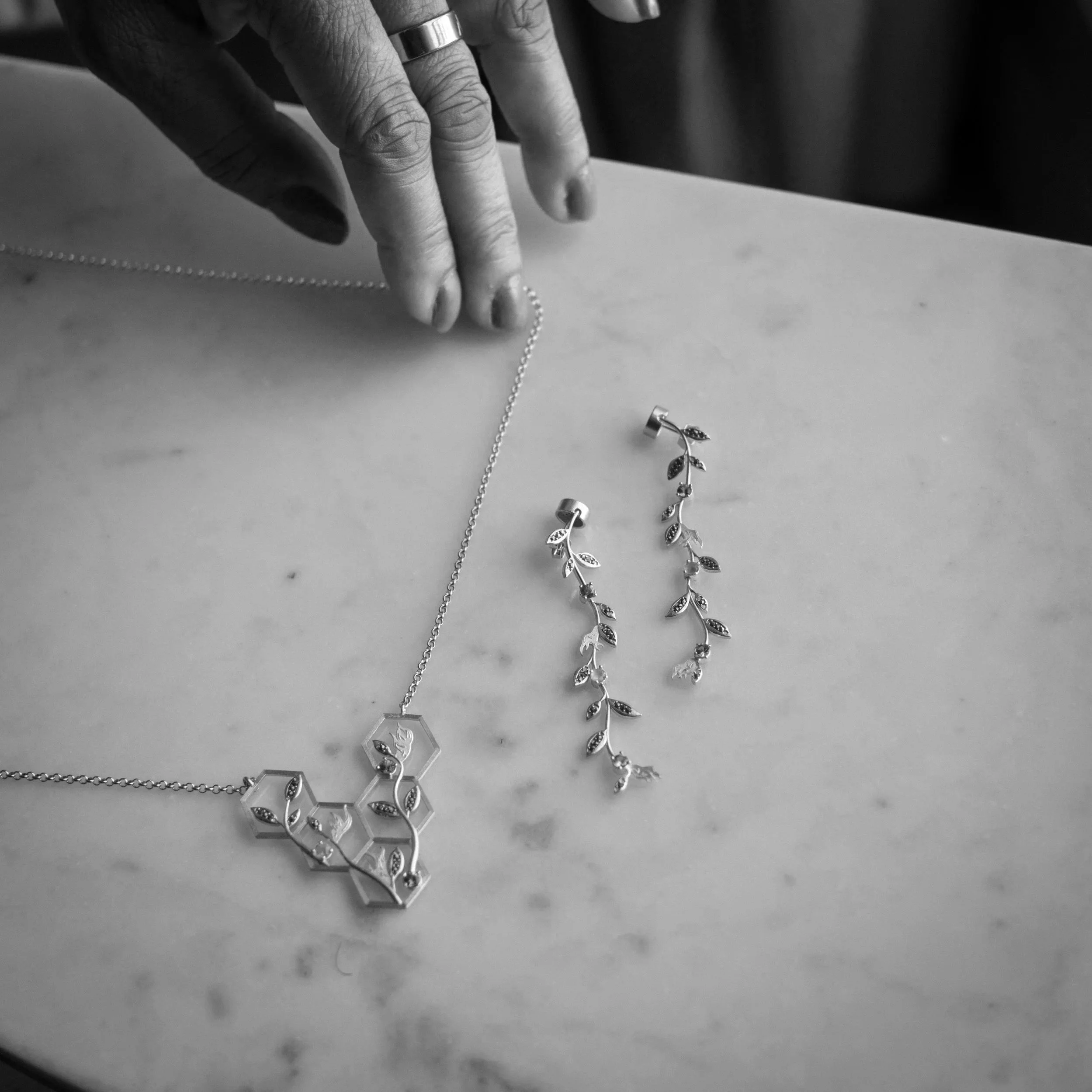 Close-up of a person's hand wearing a ring, reaching for a necklace on a white marble surface, with other jewelry including earrings and a bracelet nearby.