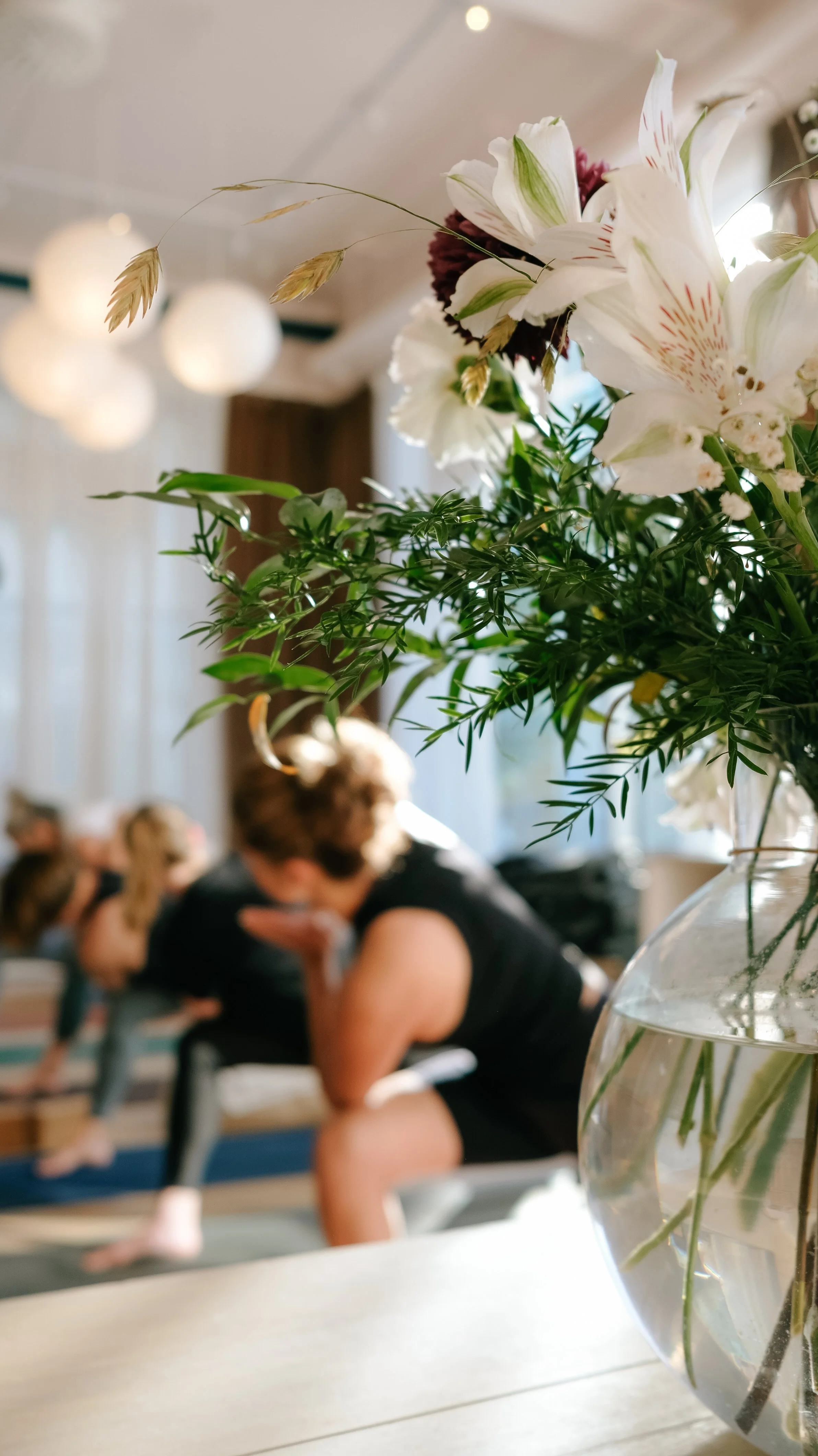 Close-up of a clear glass vase with white lilies, green leaves, and beige grass stems in the foreground. Blurred people are seated and talking in the background, suggesting a social gathering or event.