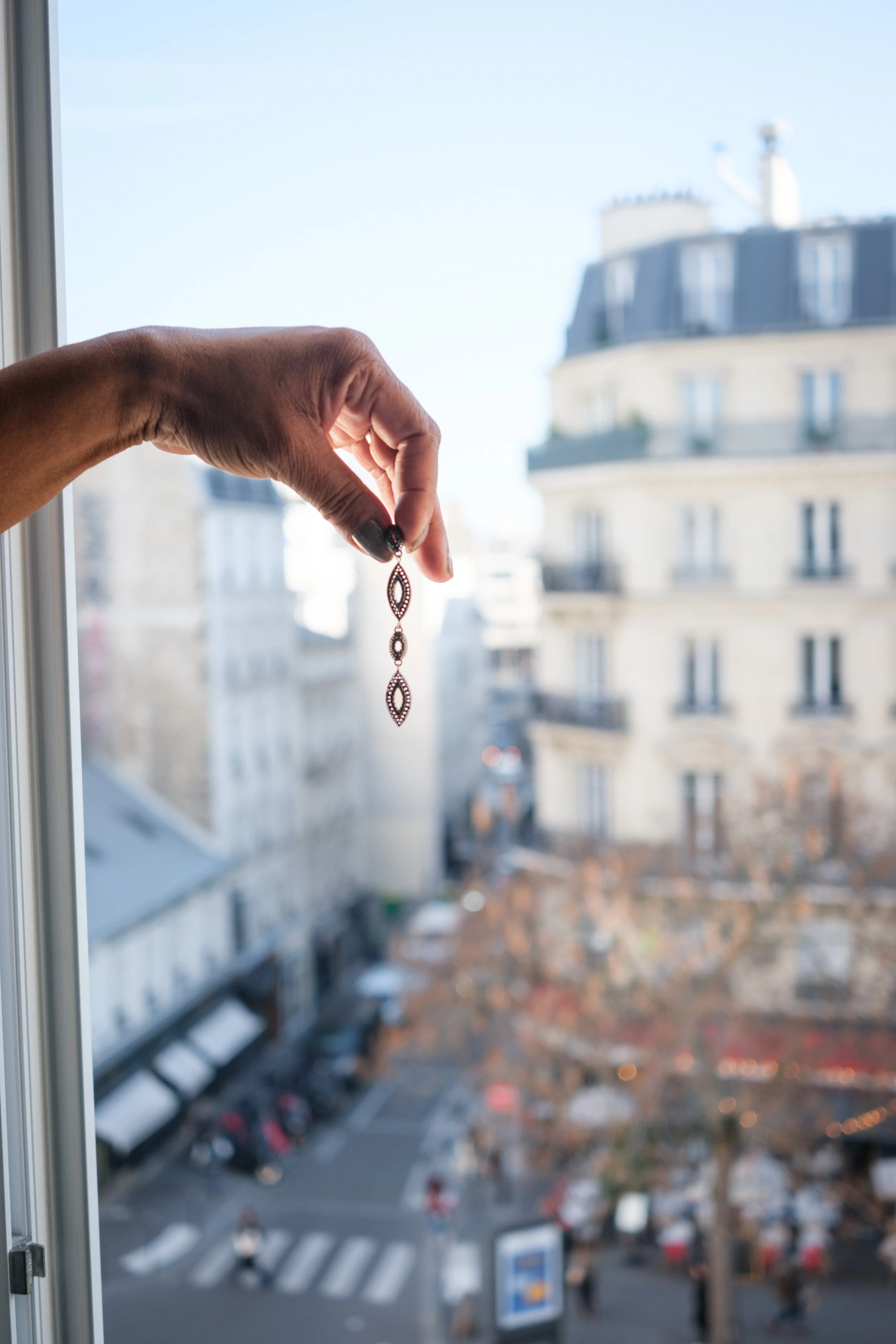 A person holding a delicate, ornate necklace near a window with a cityscape and building rooftops in the background.