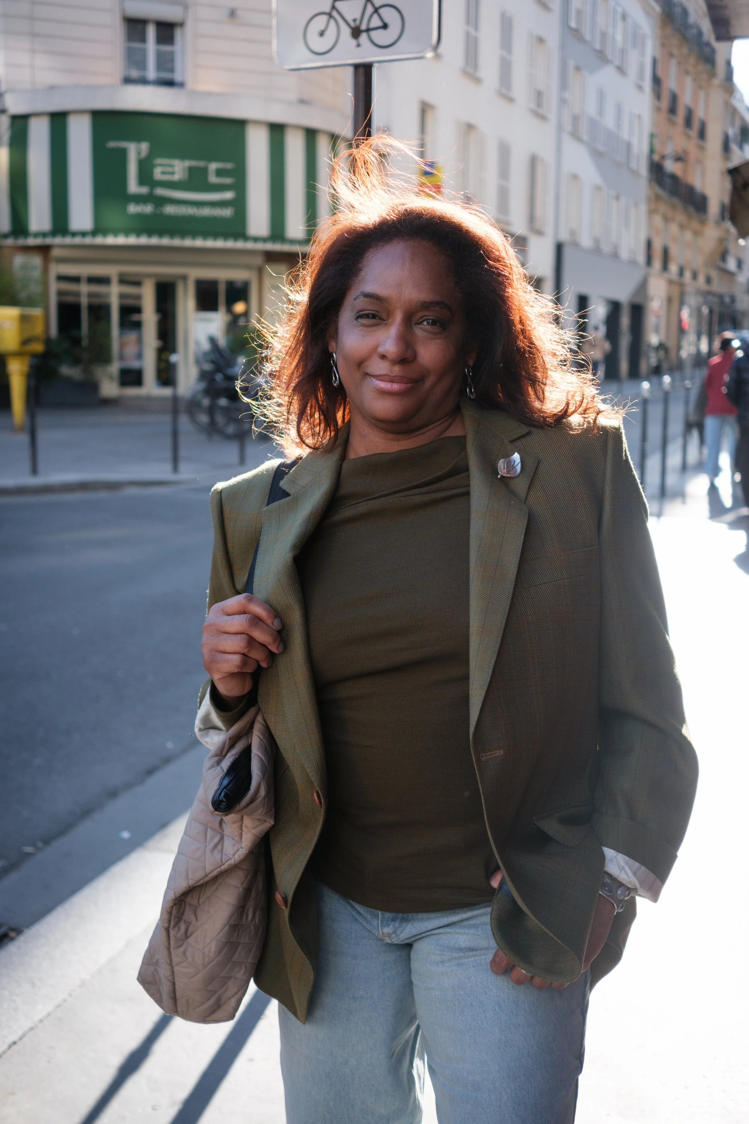 A woman walking on a city street with buildings and a sign that says 'L'arc' in the background, sunlight behind her, holding a quilted jacket over her arm and carrying a handbag.