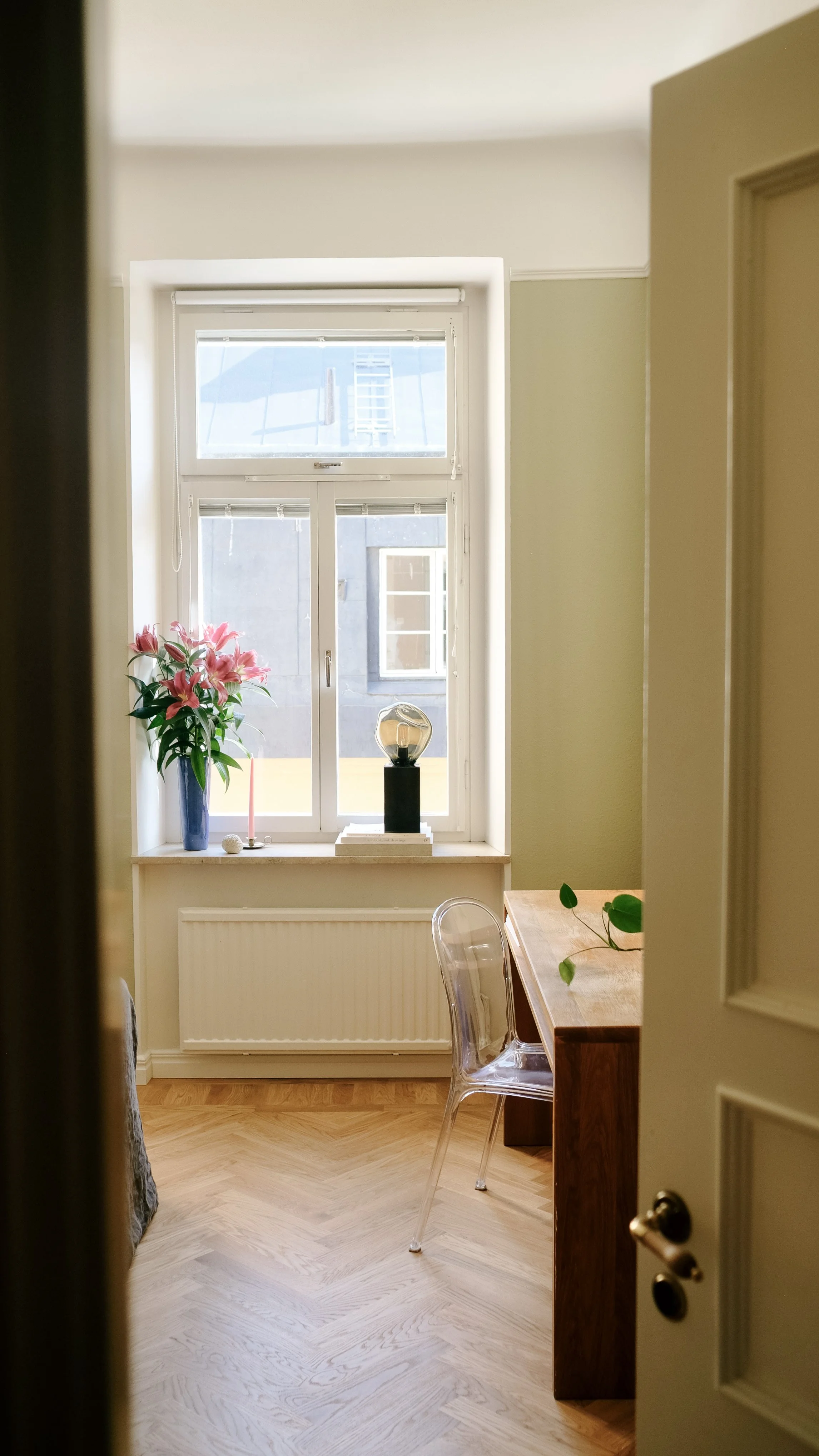 A cozy room with a large front window, decorated with pink lilies in a tall blue vase, a pink candle, a decorative object, and books. Part of a wooden table and transparent chair are visible, with sunlight illuminating the space.