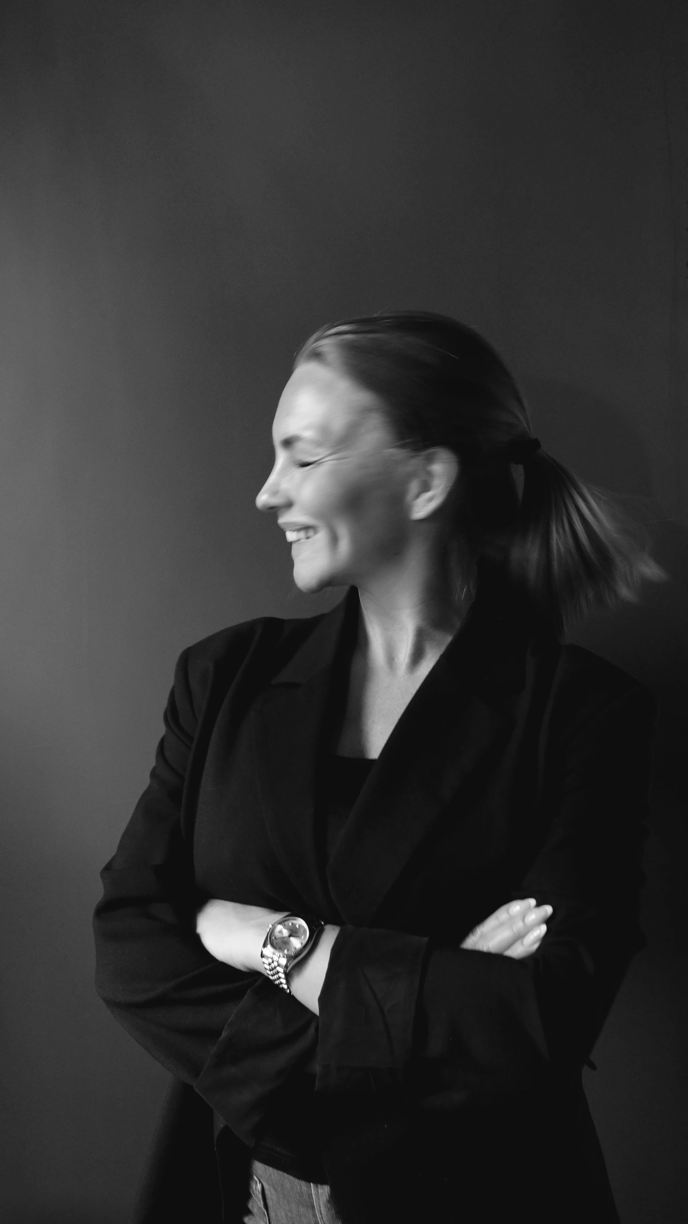 A woman laughing with her eyes closed, wearing a black blazer and a watch, with her arms crossed, against a plain dark background.