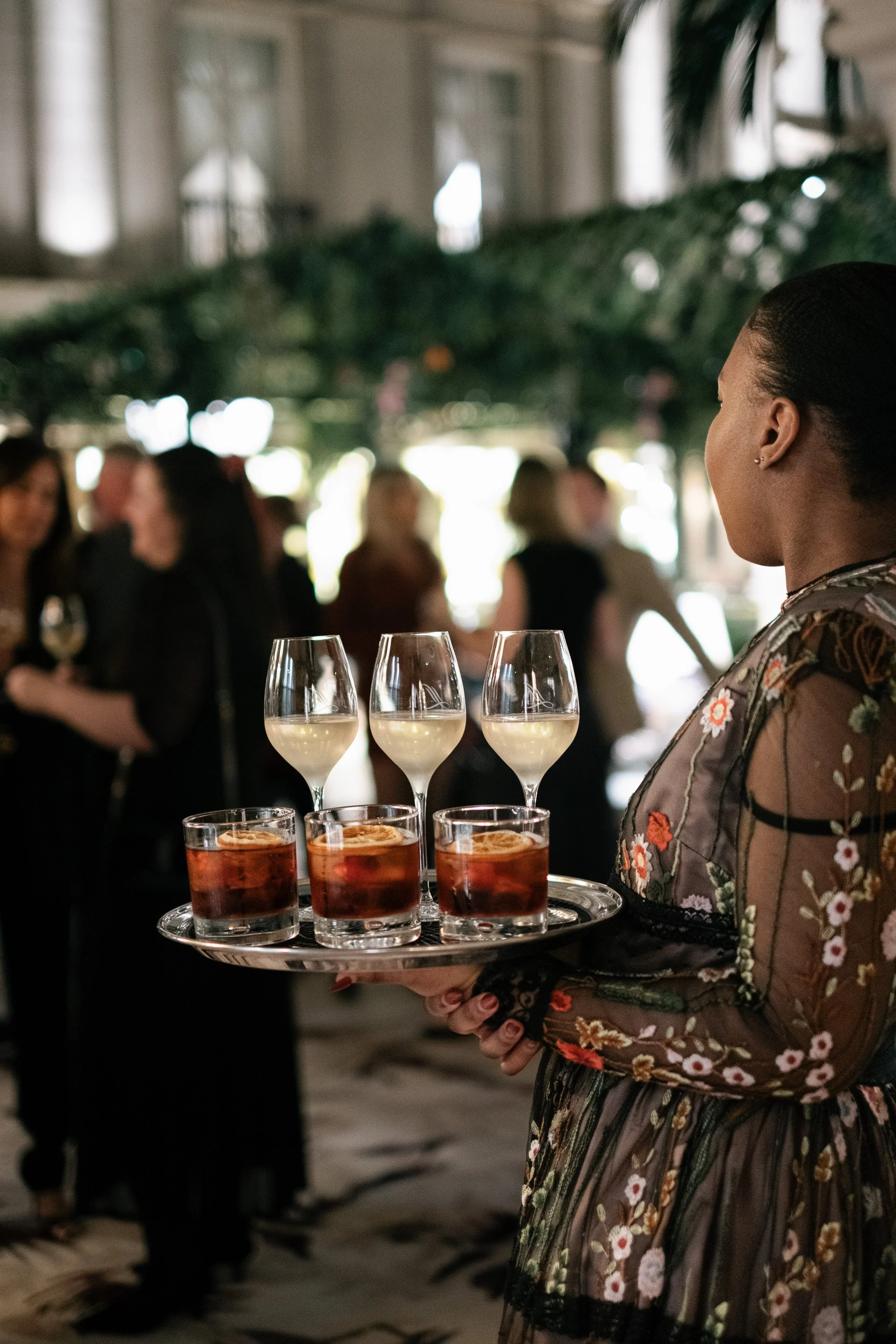 A woman dressed in floral attire holding a tray with three glasses of champagne and four glasses of a red cocktail at a social event.