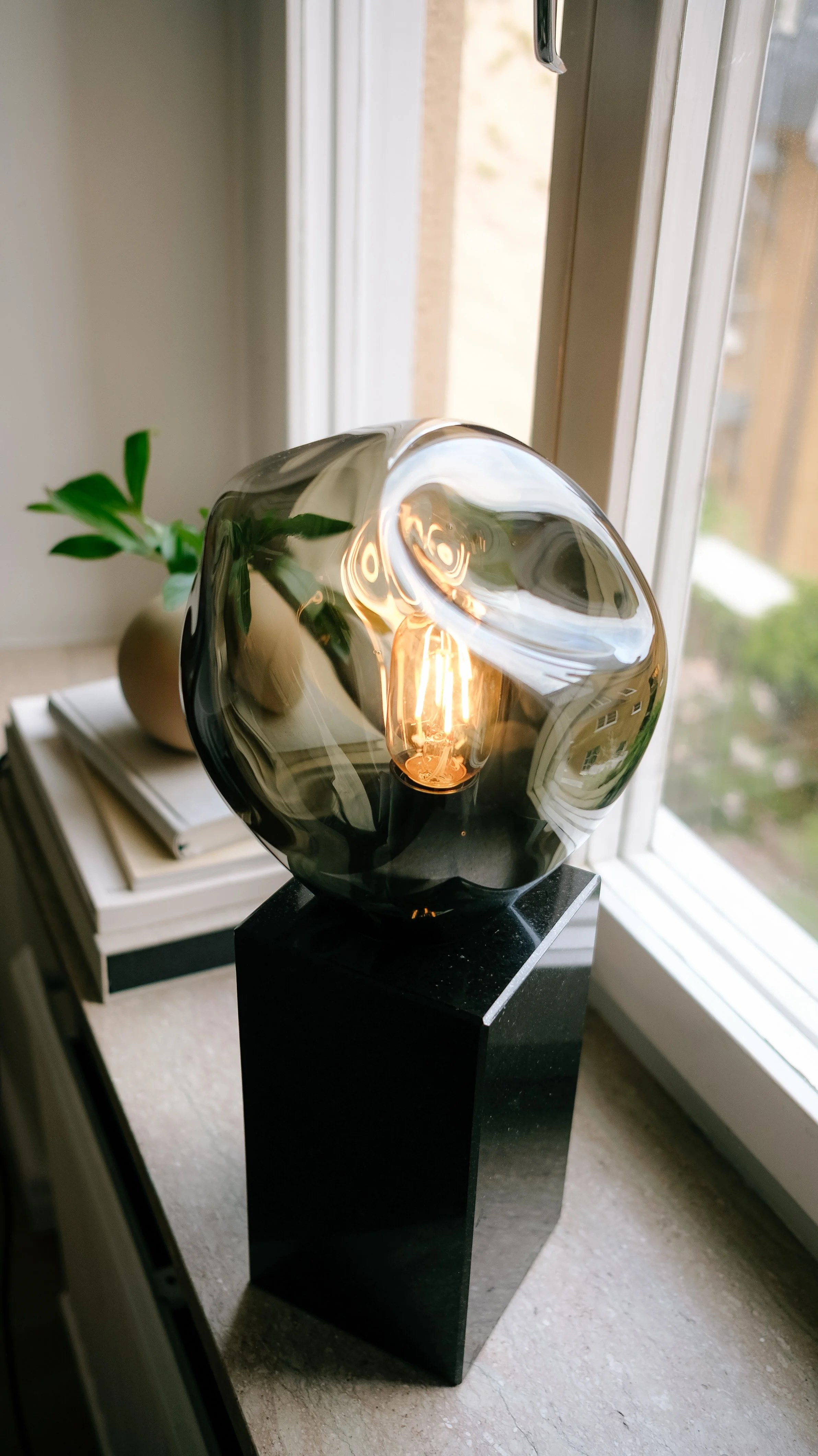 Decorative black and gold glass sculpture with an illuminated filament bulb inside, placed on a black pedestal near a window with a plant and books in the background.